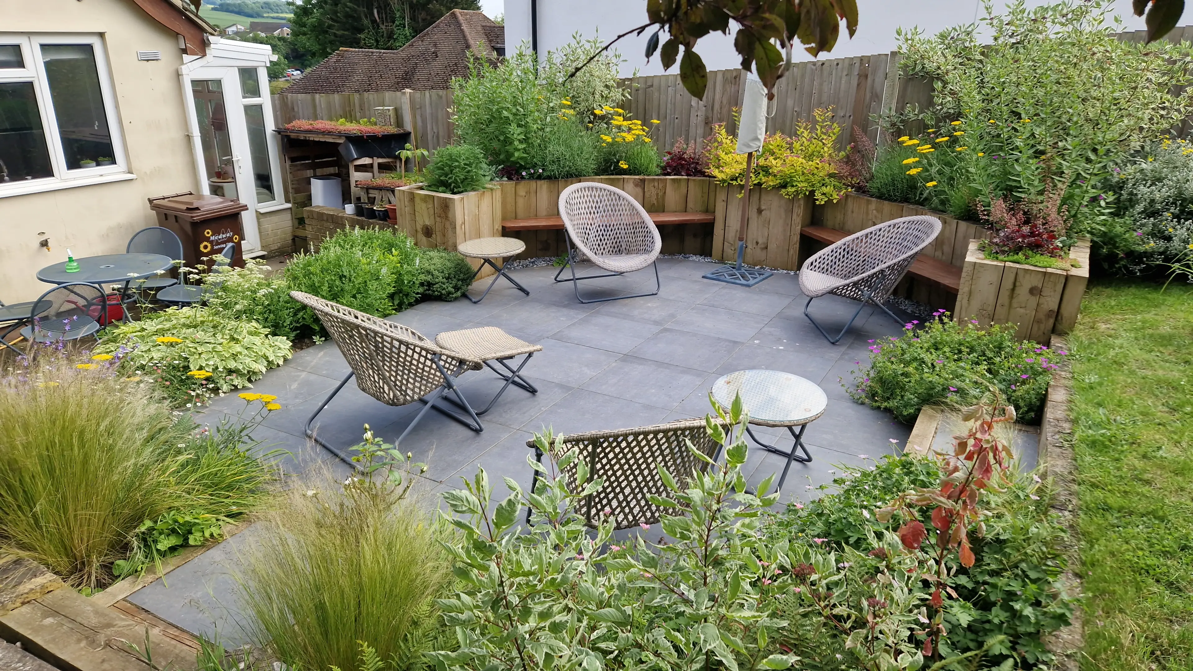 A serene garden featuring grey paving, lush greenery, and comfortable seating areas for relaxation.