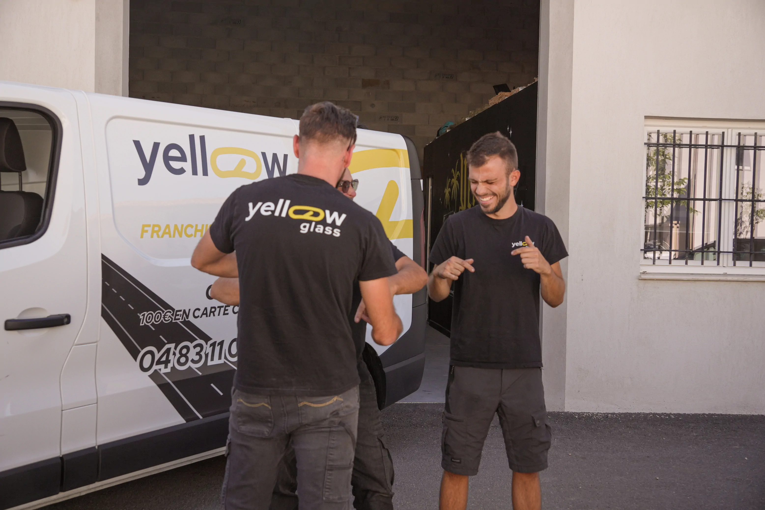 Trois hommes devant un camion jaune, l'un montre quelque chose à un autre, souriant, avec le logo de l'entreprise. 