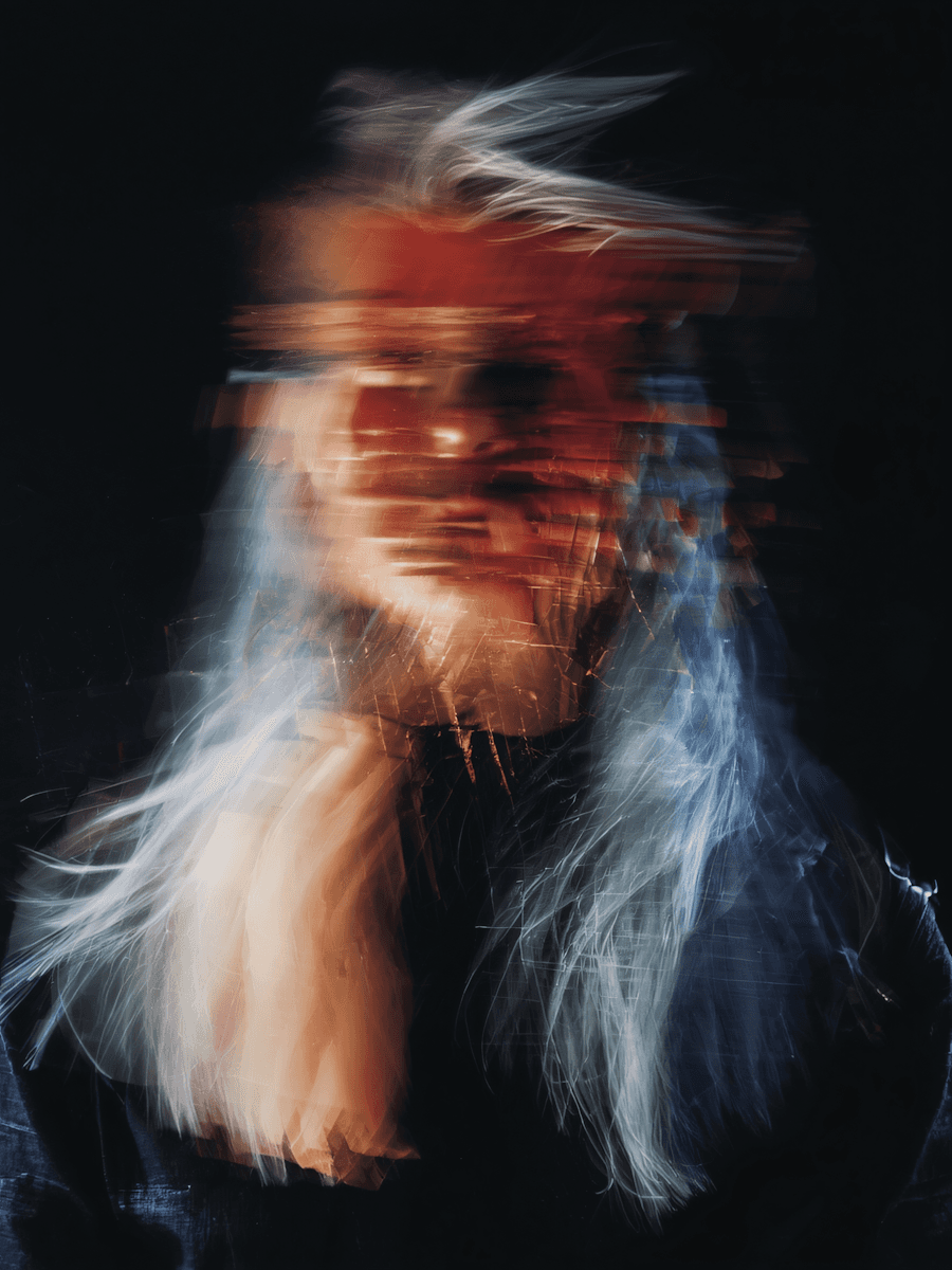 Abstract portrait of a person with long hair, captured in motion with blurred light and color streaks creating a surreal, distorted effect against a dark background.