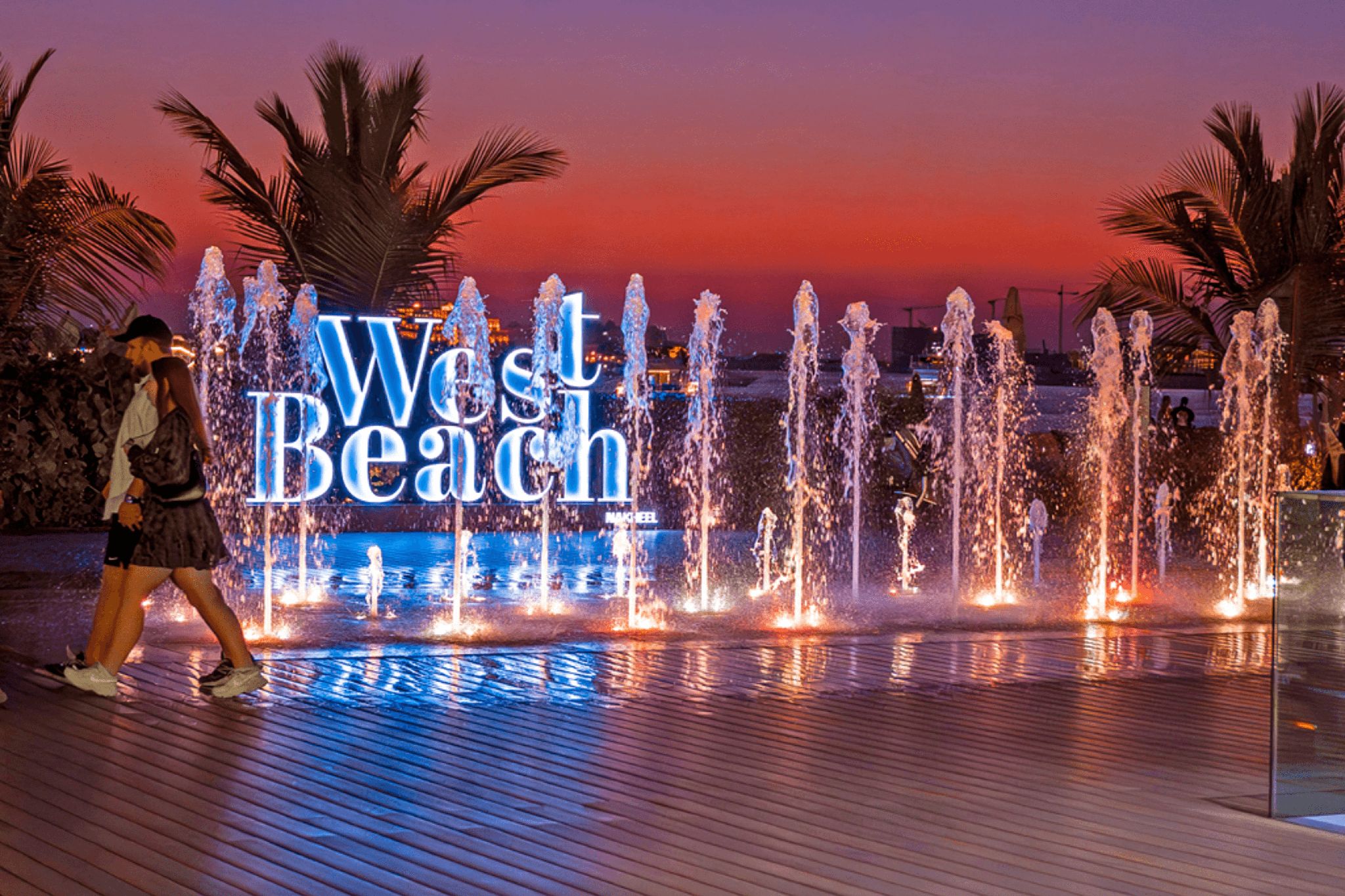 A colorful fountain show at the Palm West Beach. There are two people walking along.