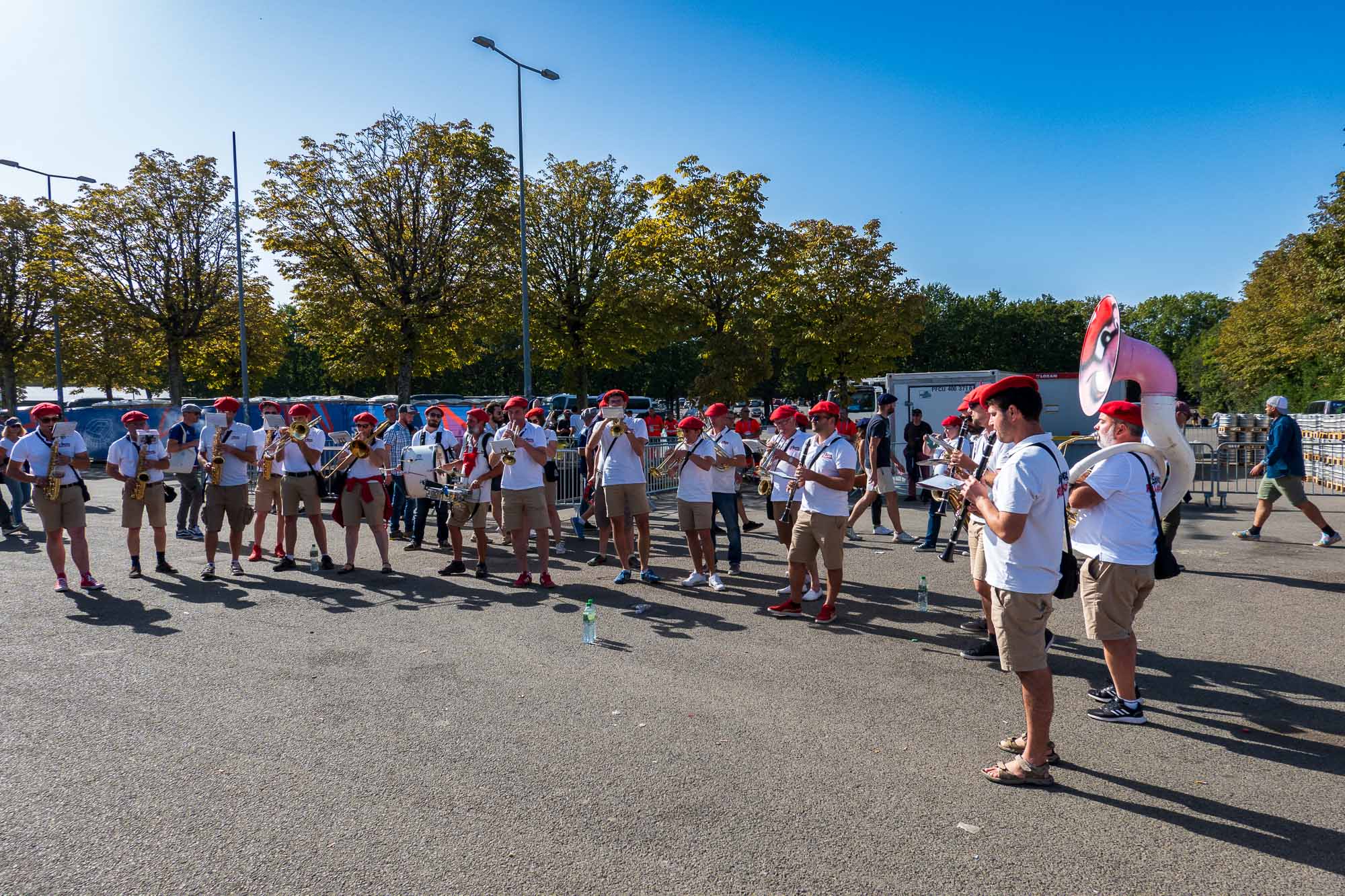 Fanfare en bérets rouges et polos blancs jouant sur un parking, sousaphone rose au premier plan, arbres et ciel bleu