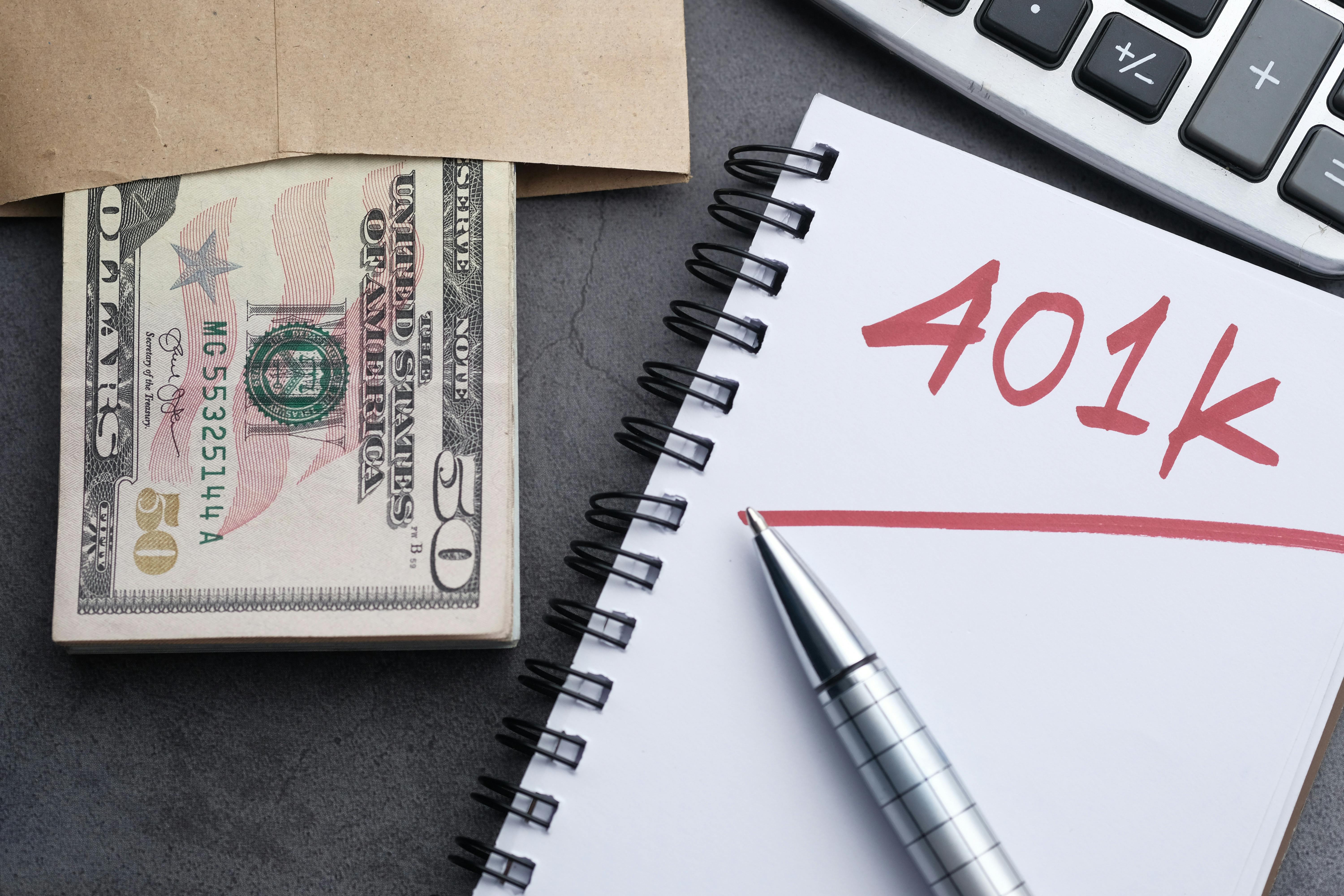 A notebook with '401K' written in bold red letters, placed beside a stack of fifty-dollar bills in an envelope, a pen, and a calculator, symbolizing retirement savings and financial planning.