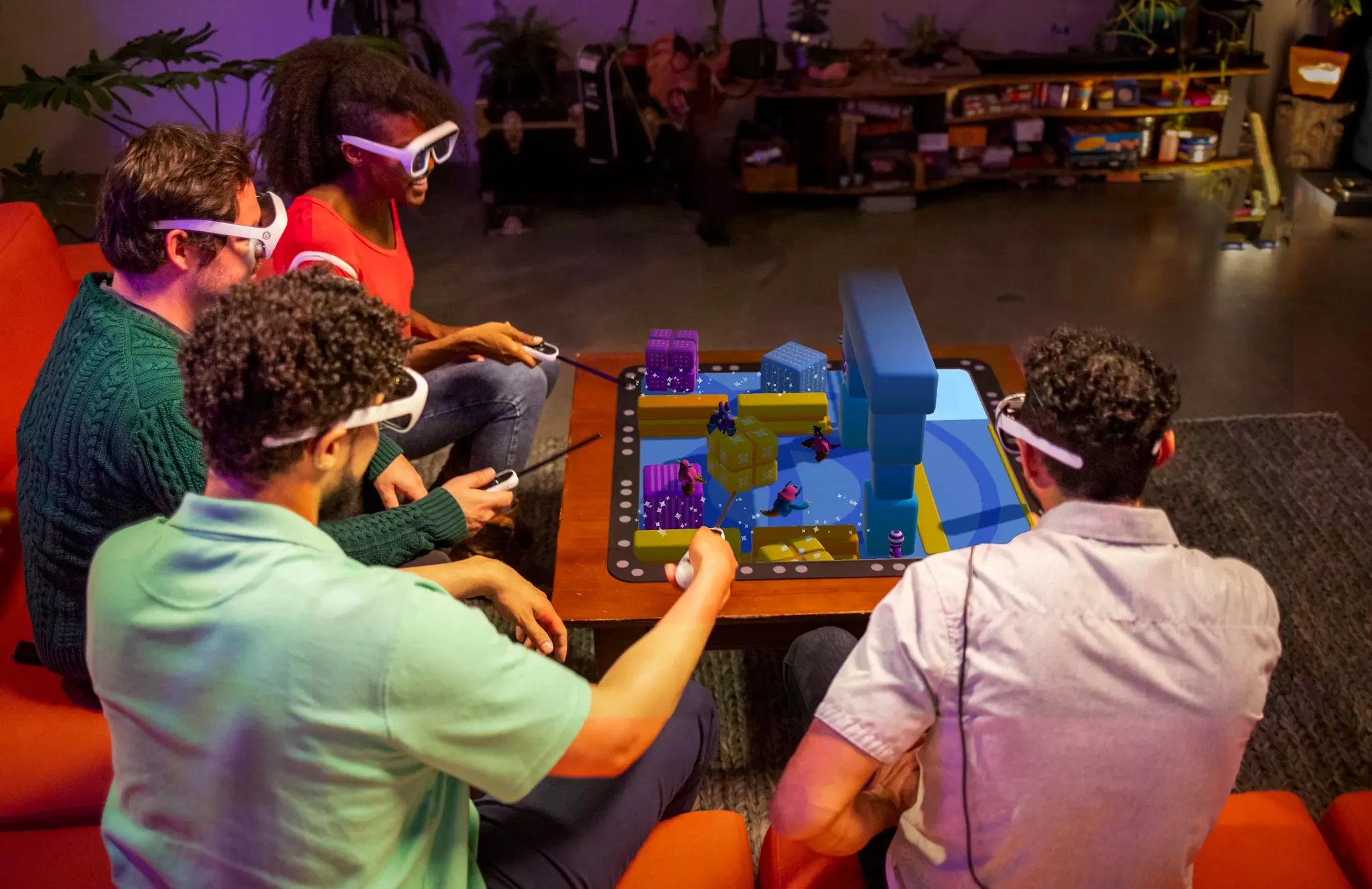 Four people wearing Tilt Five glasses use handheld controllers to play an AR tabletop game with colorful blocks and characters.