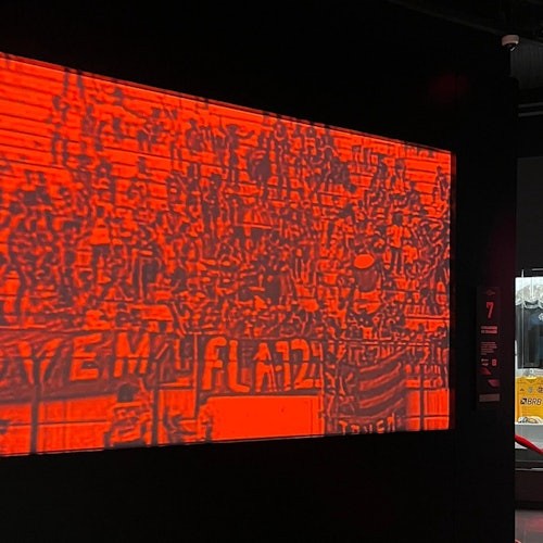 在一个黑暗房间里，一面带有红色滤镜的屏幕显示着一个模糊的群众场景。这个场景包含无法辨认的人物和文字。