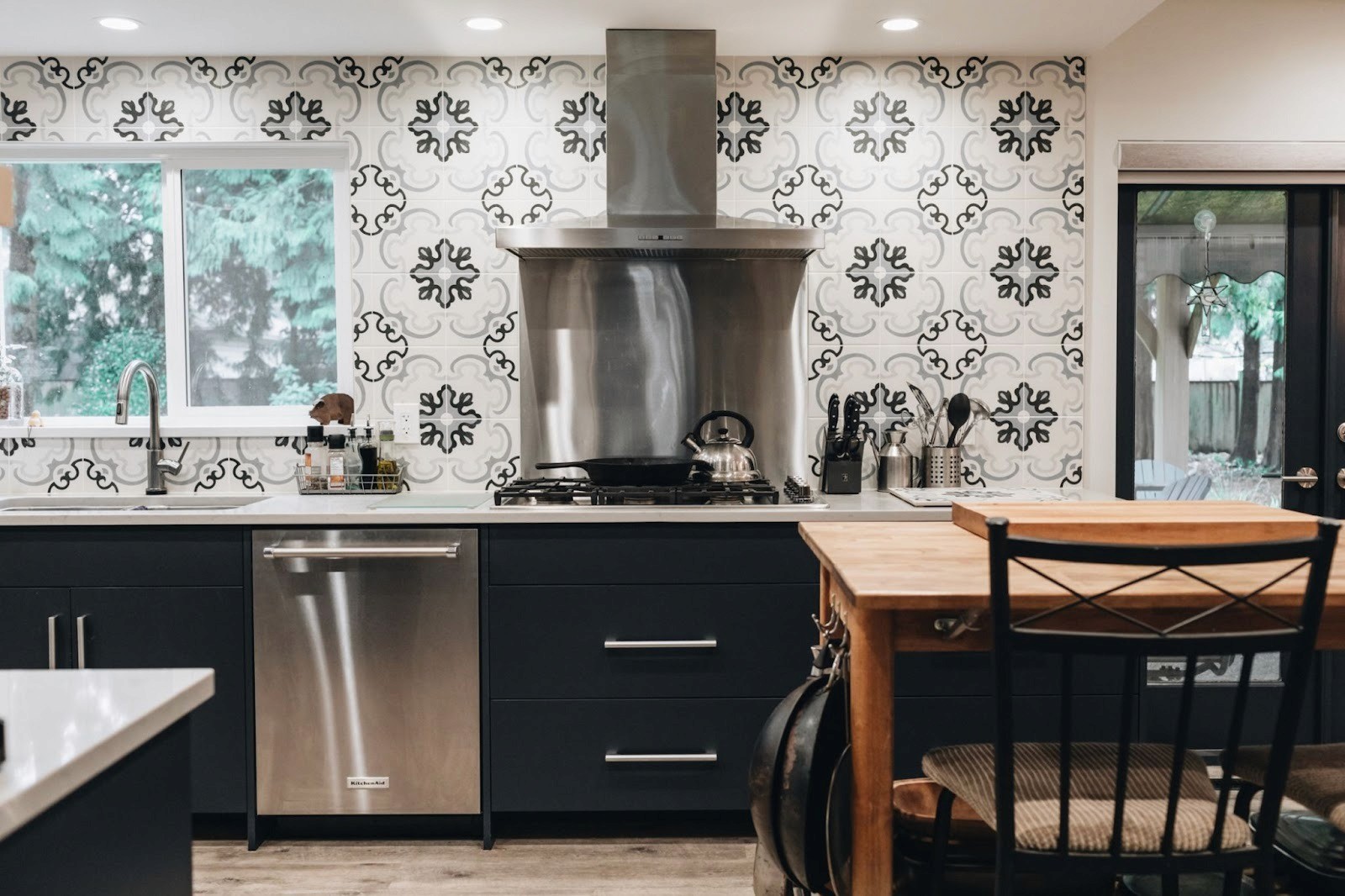 Modern kitchen with patterned tile backsplash, stainless steel appliances, dark cabinetry, and a wooden island table.