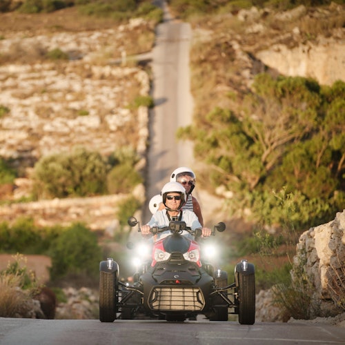 Two people wearing helmets ride a go-kart on a countryside road, surrounded by rocky terrain and greenery.