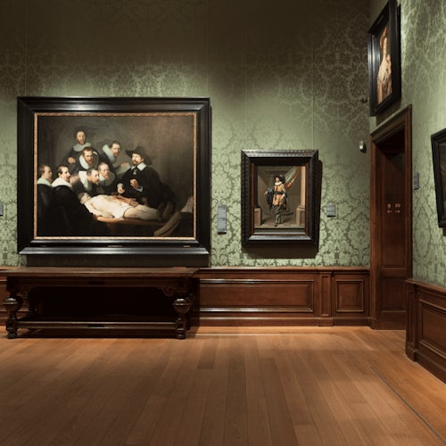 一個博物館的房間，裡面有掛在質感綠色牆上的畫框，一幅畫下面有一張木凳子，還有硬木地板。