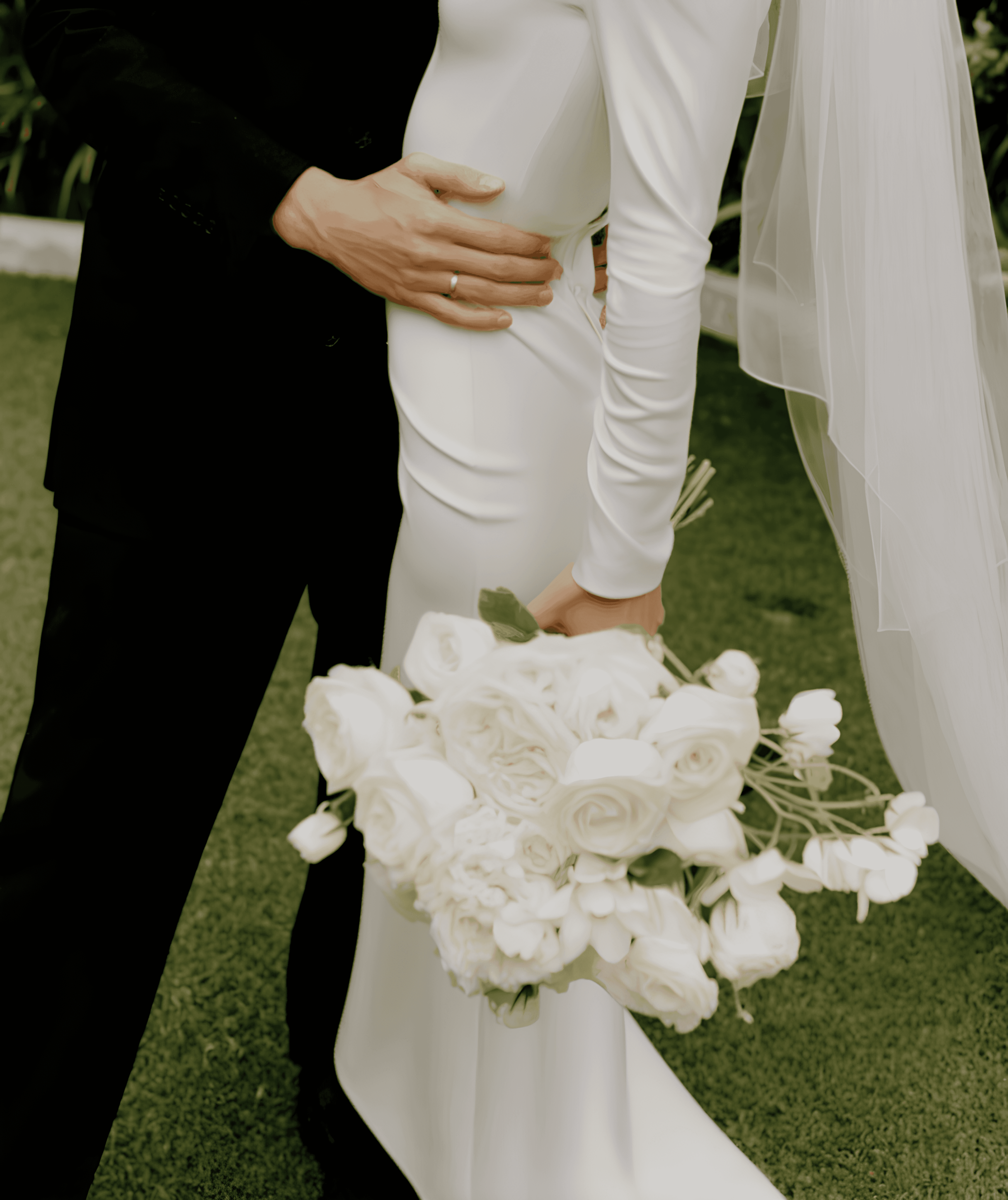 Close-up of a bride and groom standing close together outdoors; the groom in a black suit gently rests his hand on the bride’s pregnant belly over her sleek white long-sleeve wedding gown, while she holds a lush bouquet of white roses; her veil flows softly in the background.