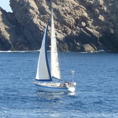 A sailboat with white sails and several people onboard sailing near rocky cliffs in a calm blue sea.