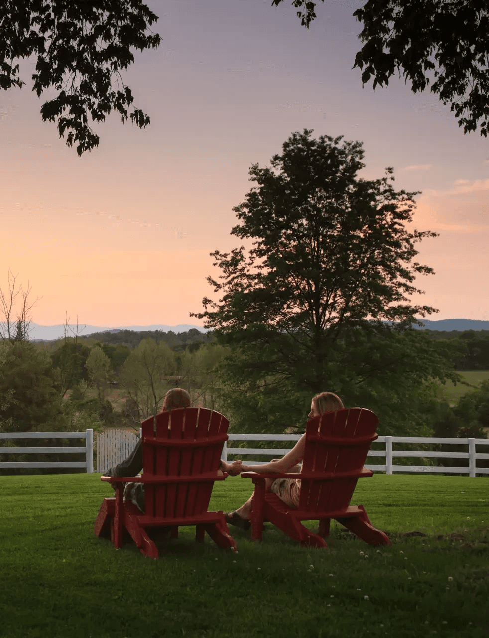 Two people sit in red Adirondack chairs on a lush green lawn, facing a serene sunset view with trees and rolling hills in the background at The Estate at Meander.
