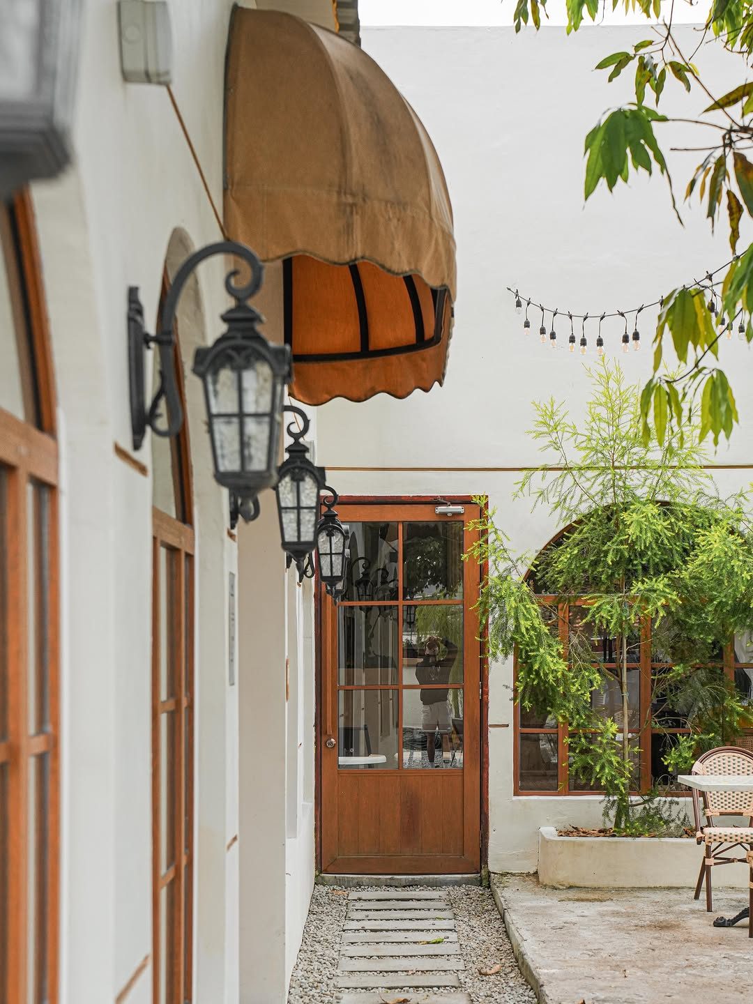 A serene pathway leading to a wooden door, flanked by white walls, decorative lanterns, and lush greenery.