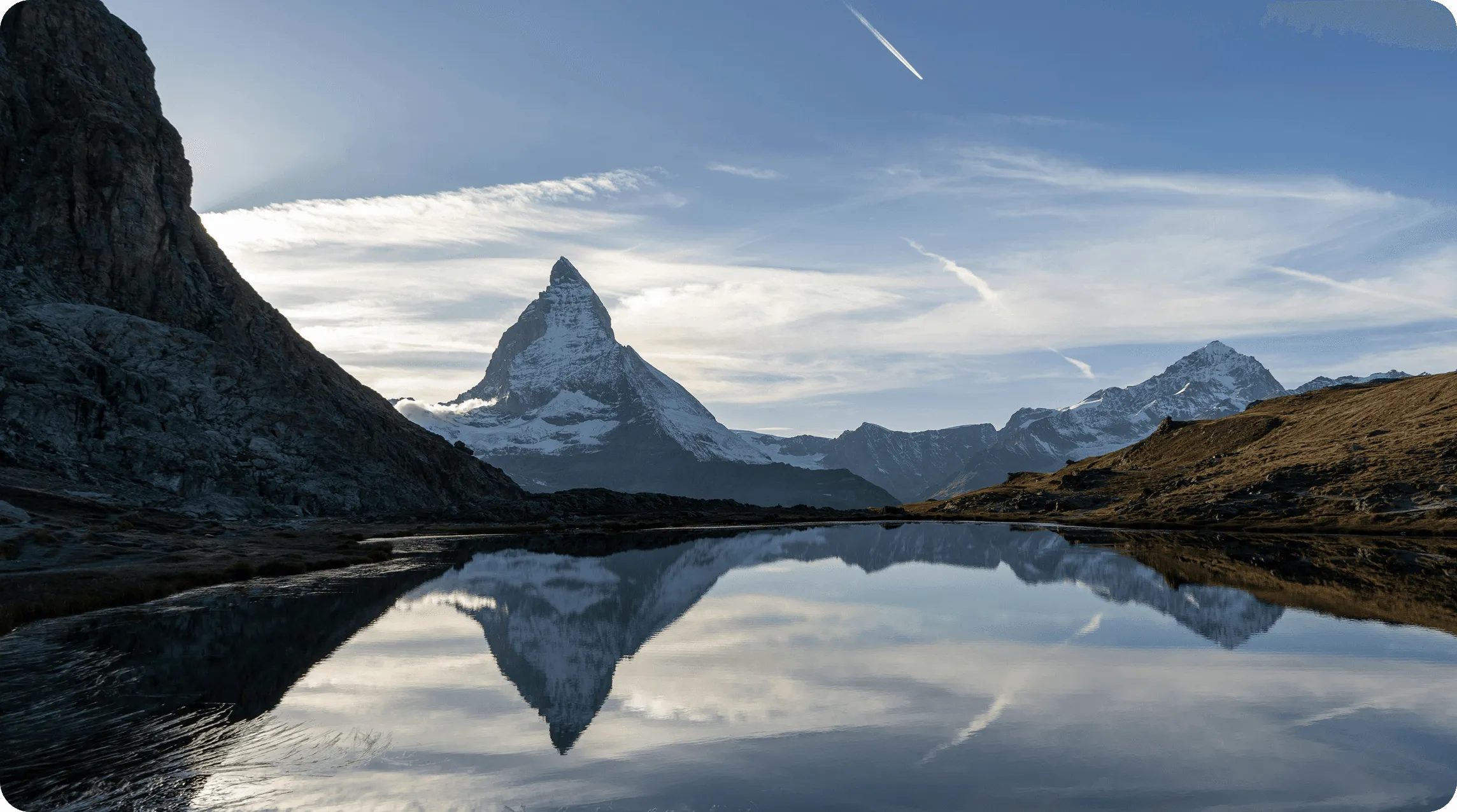 A serene mountain landscape reflecting in calm water under a clear sky, with peaks and gentle slopes surrounding.