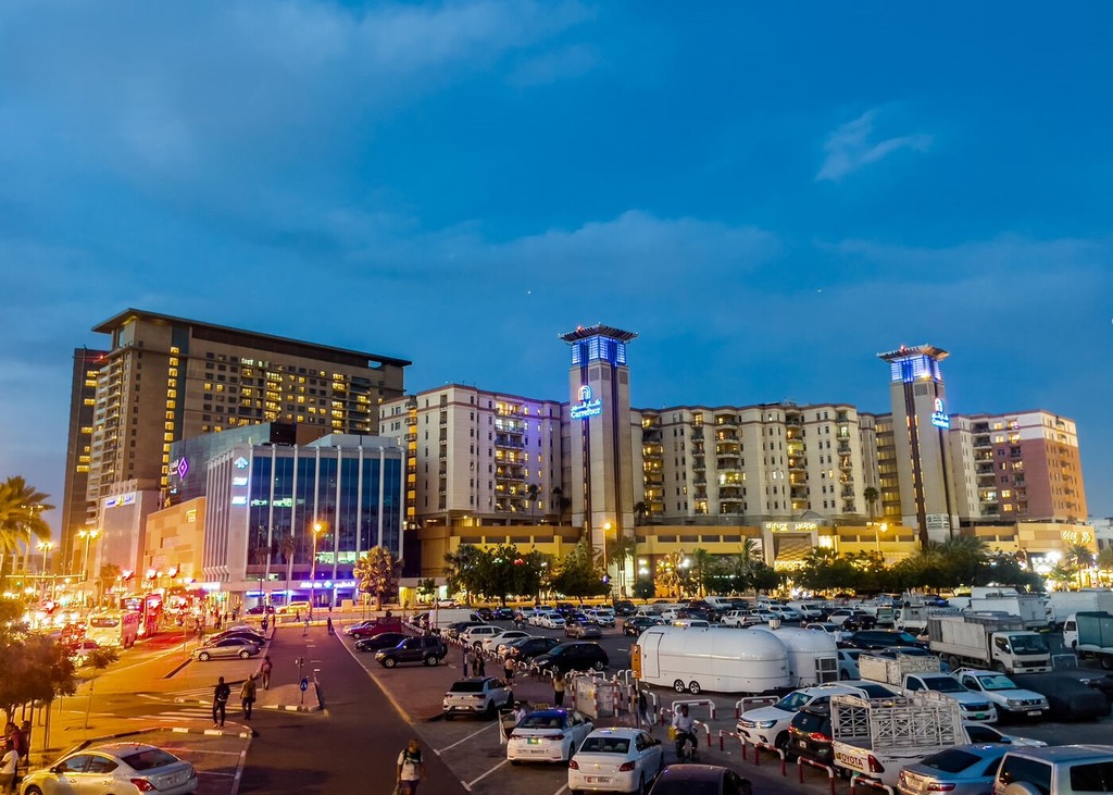Evening view of Grand Oasis hotel in Deira, Dubai, with lit signage, busy parking lot, and palm trees in a vibrant urban scene.
