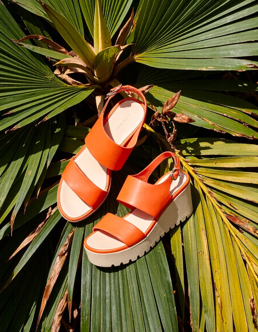Bright orange platform sandals with chunky soles arranged on green palm fronds, creating a tropical summer still life composition.