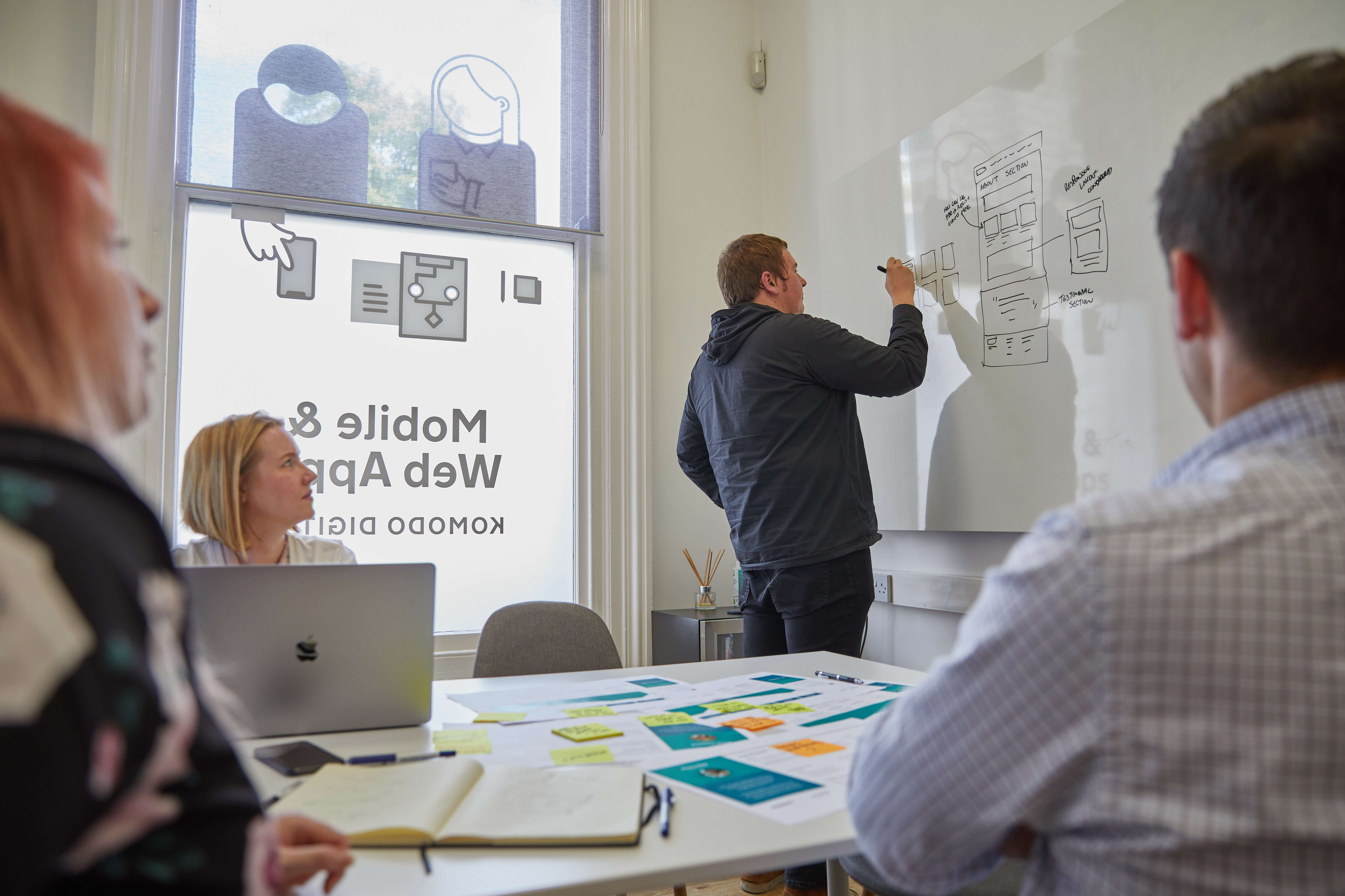 A workshop at the Komodo studio. There are post it notes on the table and one of the designers is sketching wireframes on a whiteboard