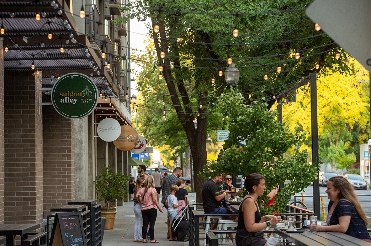 A lively urban street scene in Midtown Sacramento with people dining and socializing outdoors at various cafes and restaurants.