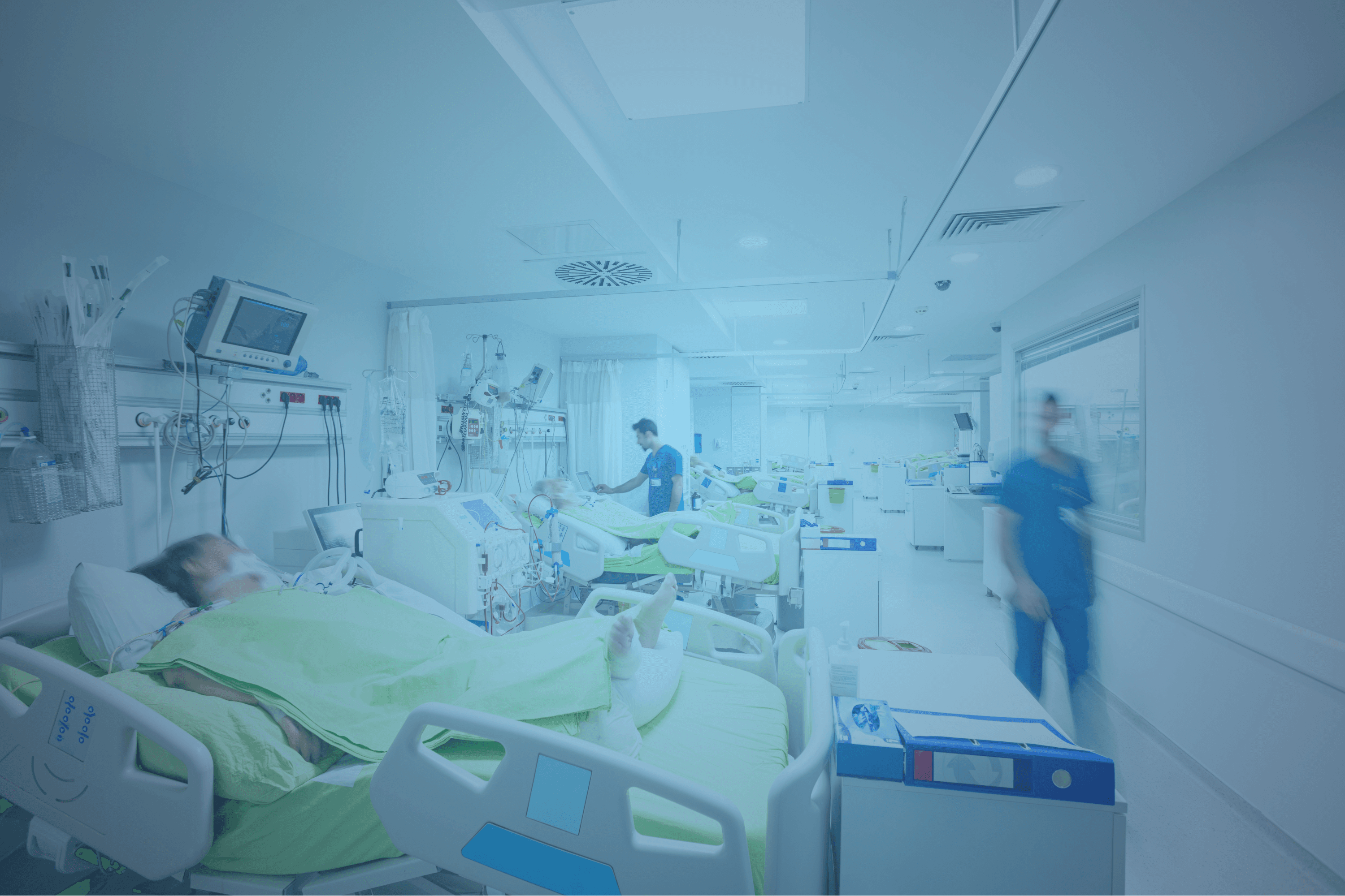 Patient liegt in einem Krankenhausbett auf der Intensivstation, überwacht von medizinischen Geräten. Im Hintergrund Pflegepersonal in blauen Kitteln.