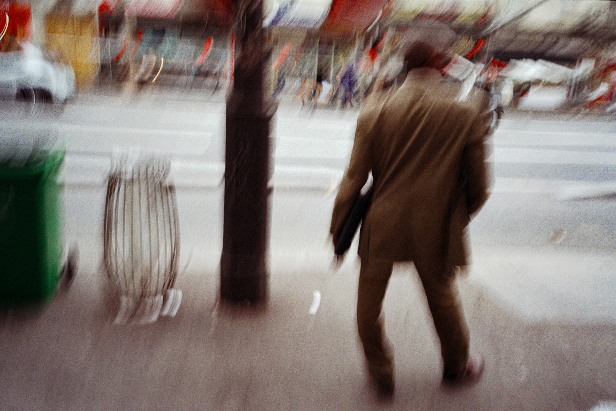 A person in motion, wearing a brown suit, walks on a busy city sidewalk with blurred cars and streetscape in the background.