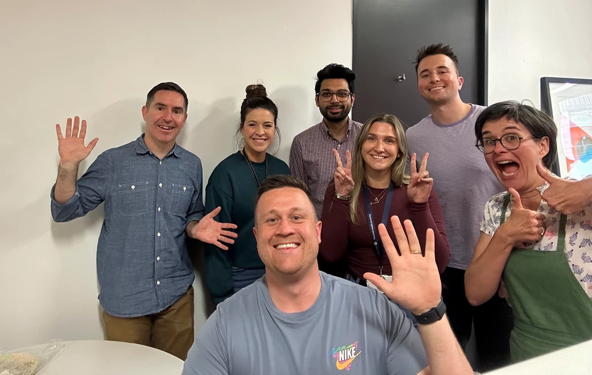 Group of seven people standing indoors, posing with raised hands and peace signs in front of a light-colored wall and door.
