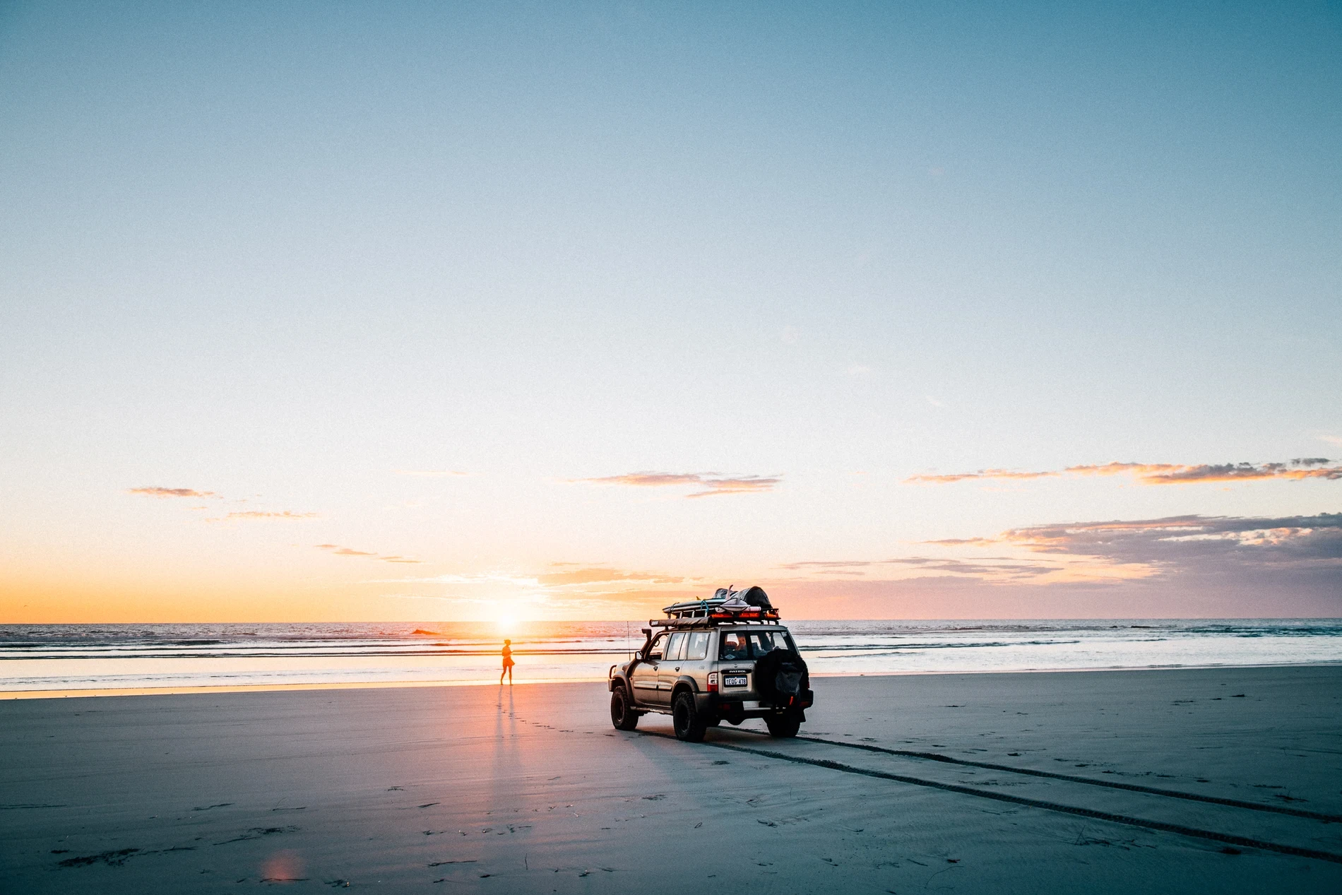 A four-wheel drive parked on wet sand at sunset, a lone figure standing in the sun's reflection