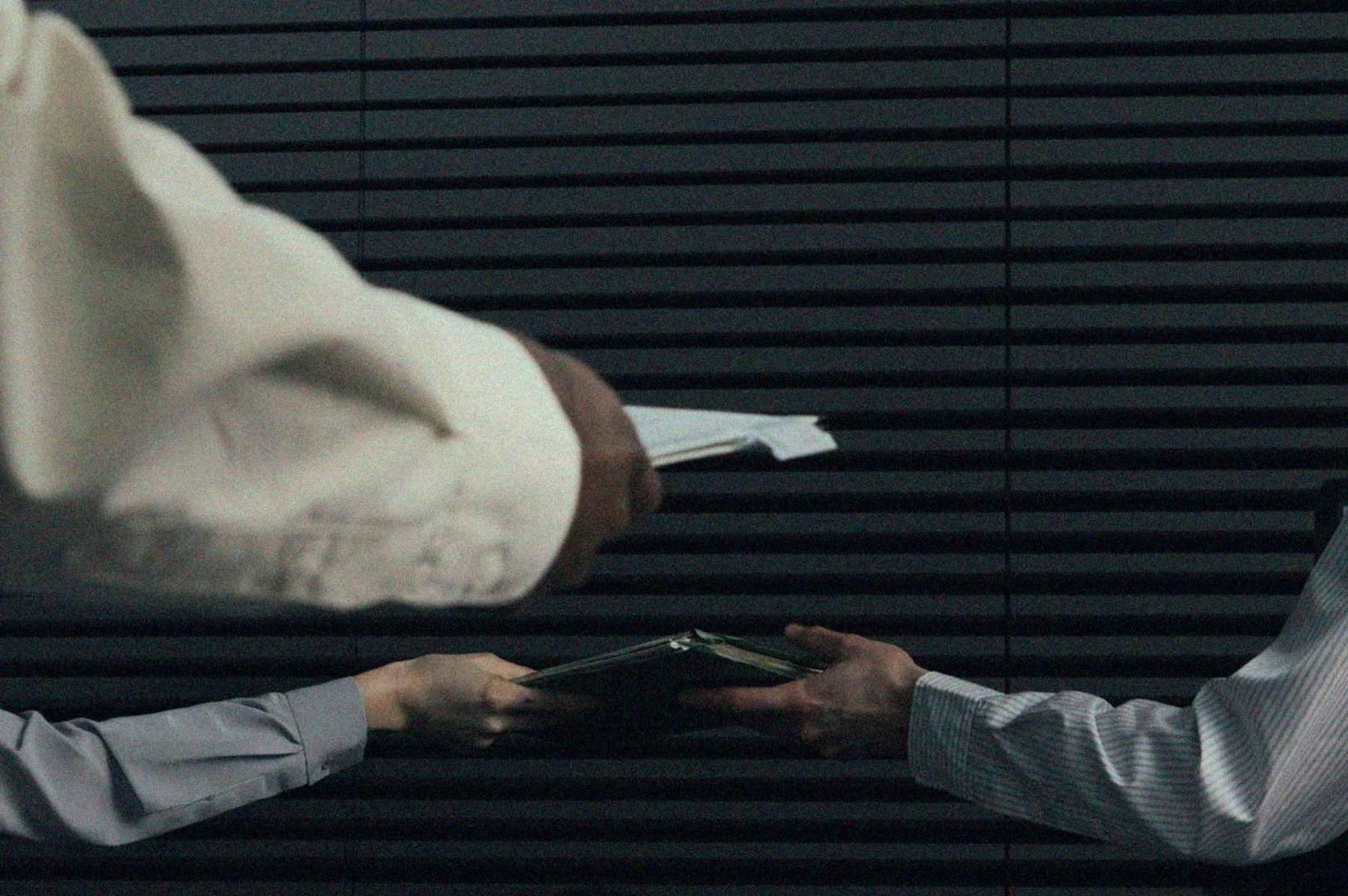 Three hands exchanging documents in a dimly lit room with horizontal blinds. The mood is tense and secretive, suggesting a confidential meeting.
