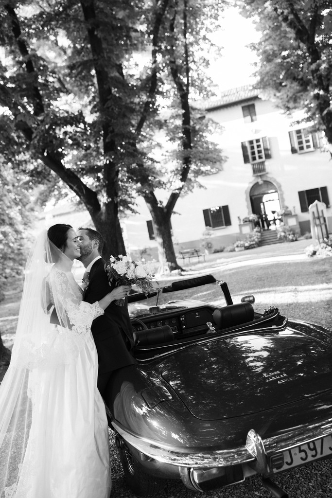 Sposi in abito bianco e nero baciano davanti a auto d'epoca sotto gli alberi della villa, reportage di matrimonio