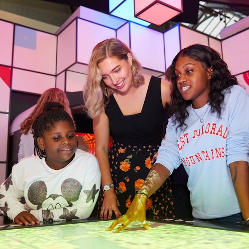 Three women, smiling and interacting with a touchscreen table, are inside a brightly lit, modern space with geometric decor.