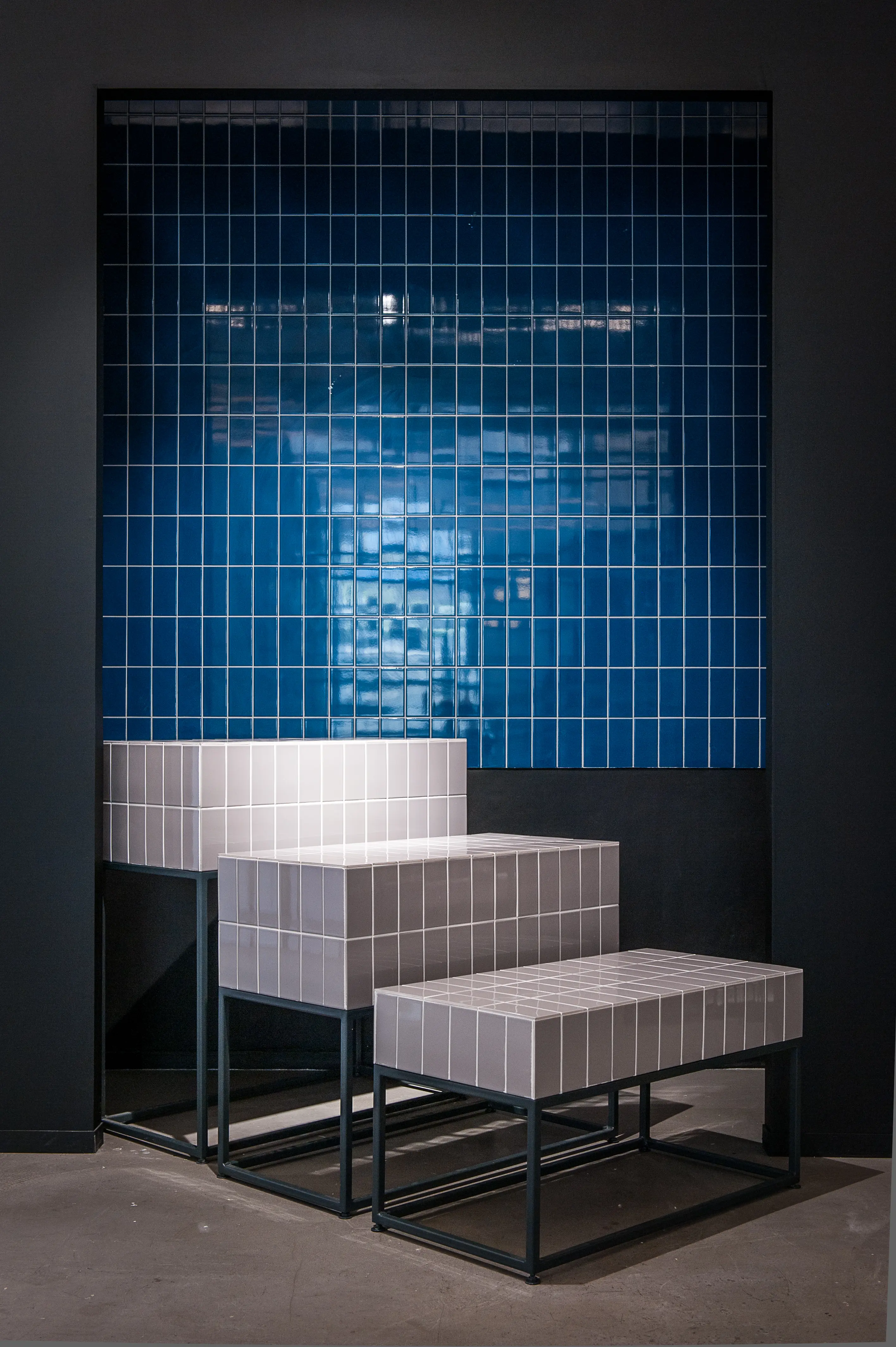 Dark niche at the 17 FASHION BRANDS showroom with a glossy blue tiled wall and stepped display pedestals covered in white tiles on black steel frames.