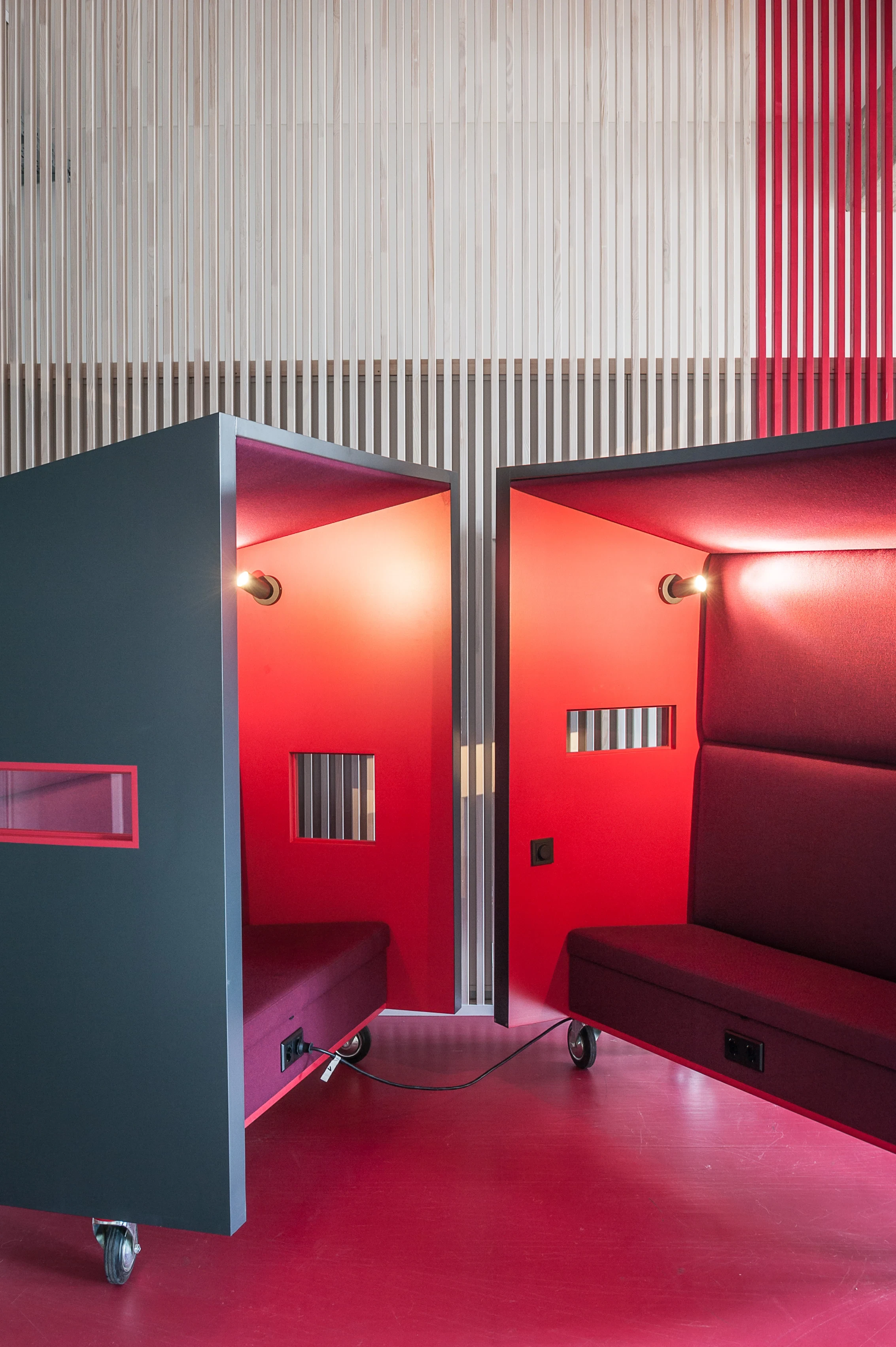 Two mobile phone-booth boxes on castors at BASILICOM, dark grey outside and bright red inside with upholstered benches and small viewing windows, in front of a striped slatted wall.