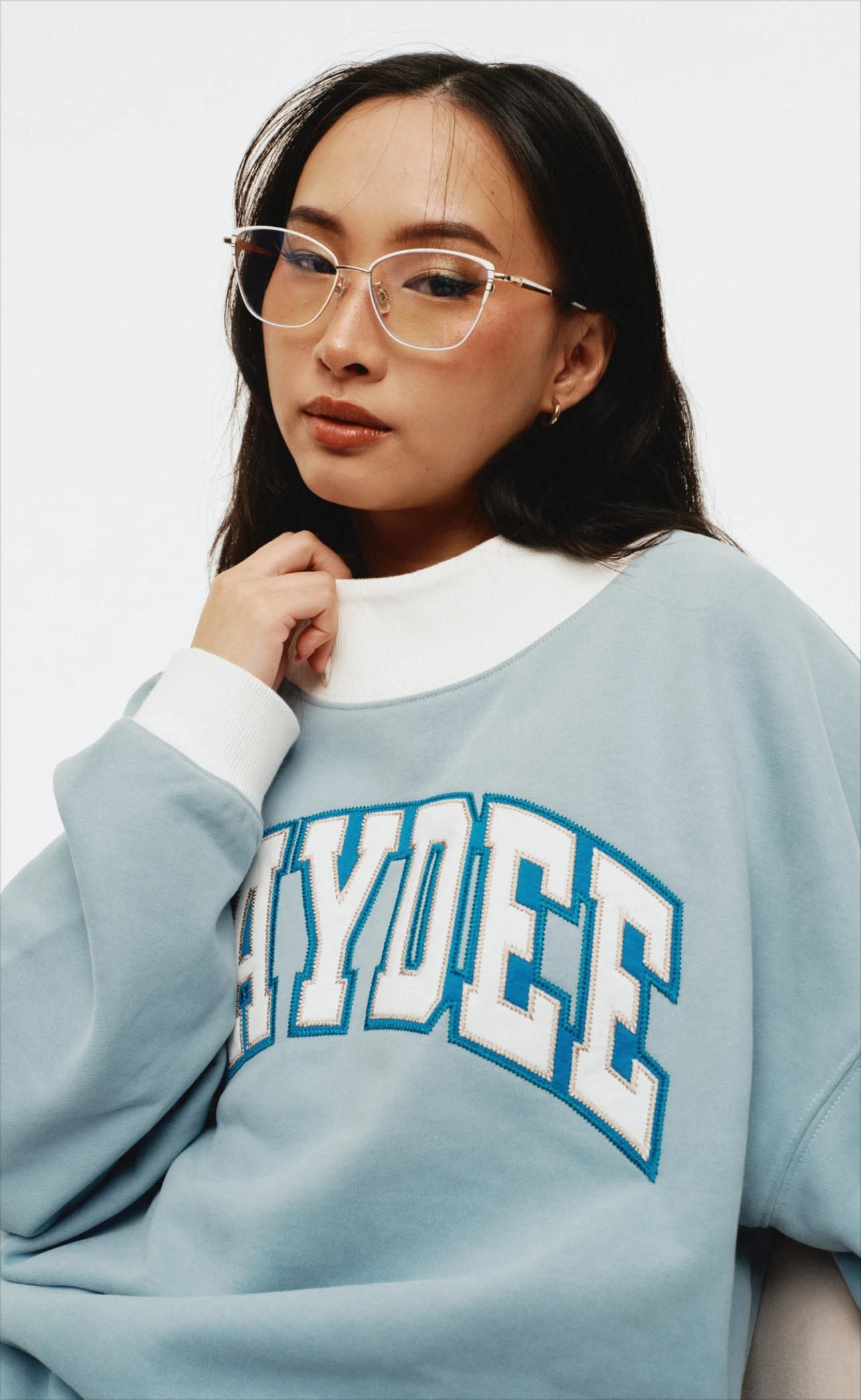 Close-up studio portrait of a model wearing a light blue “Kaydee” crewneck with a white mock-neck collar and clear-framed glasses.