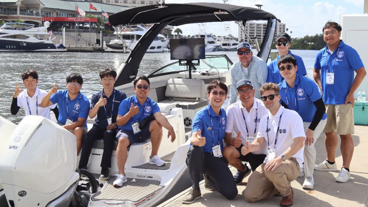 A smiling man with curly hair is sitting at a desk, looking at the camera, with a laptop and a lamp in the background.