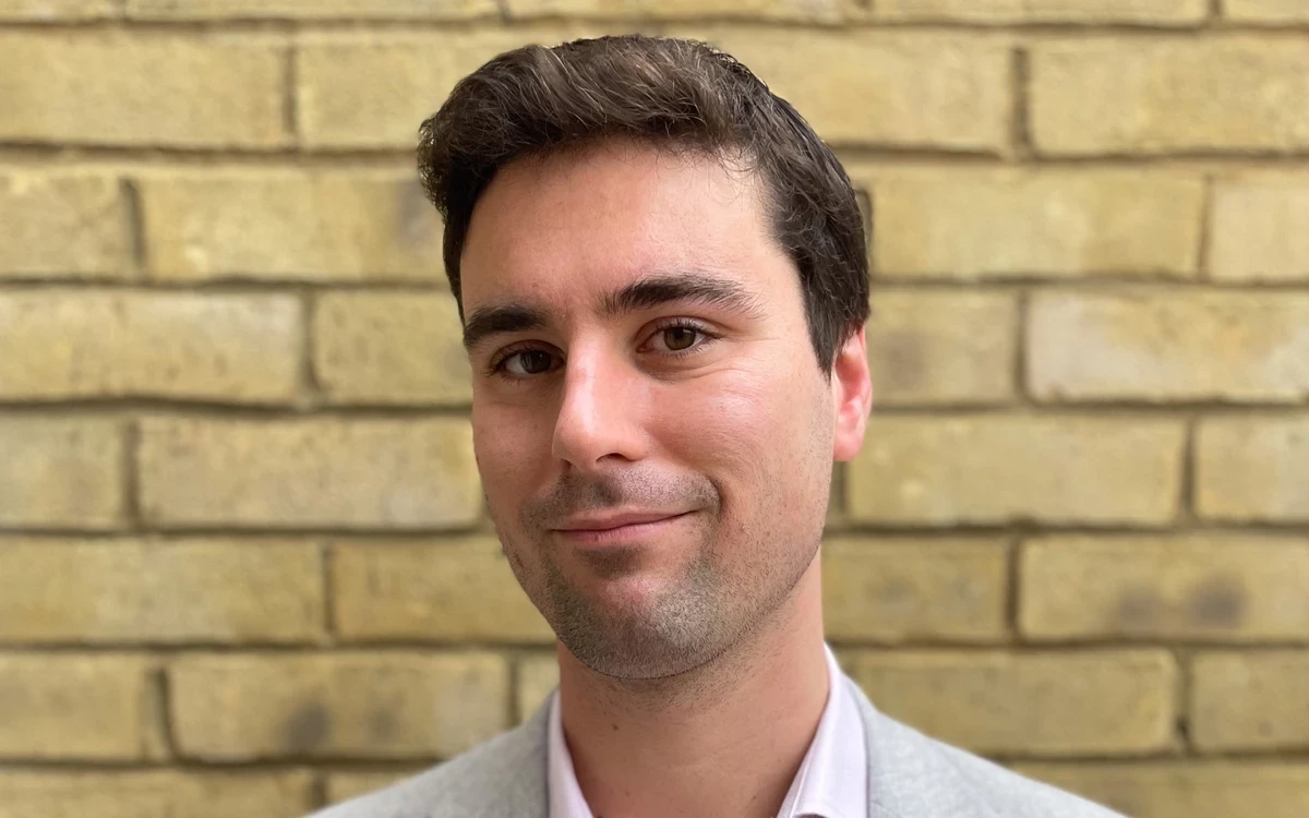 A person stands in front of a light‑coloured brick wall, wearing a light blazer and collared shirt. The photo is framed from the shoulders up.