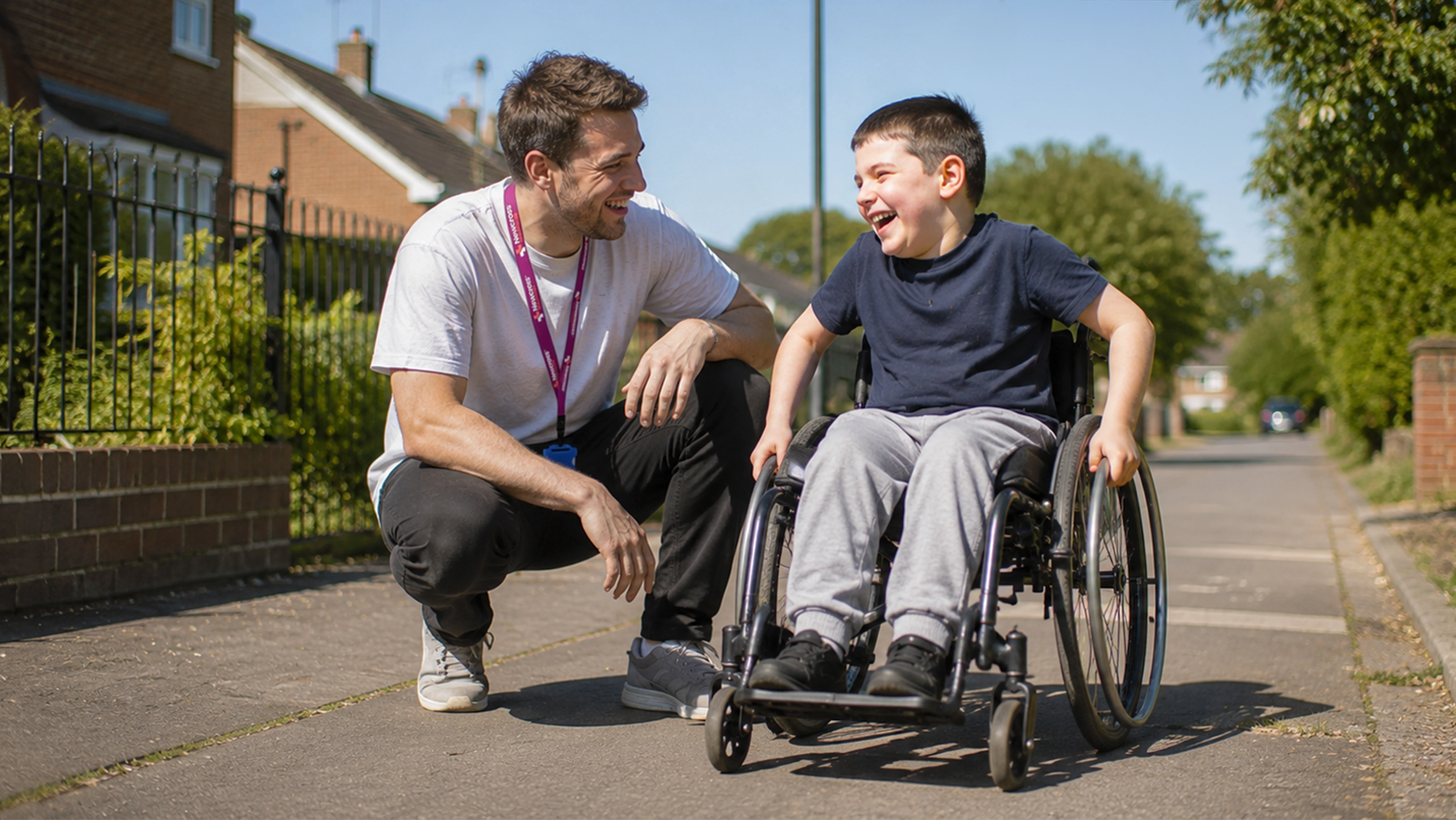 A young boy in a wheelchair and a man crouched next to him, sharing a joyful moment outdoors on a sunny day, with houses and trees in the background.