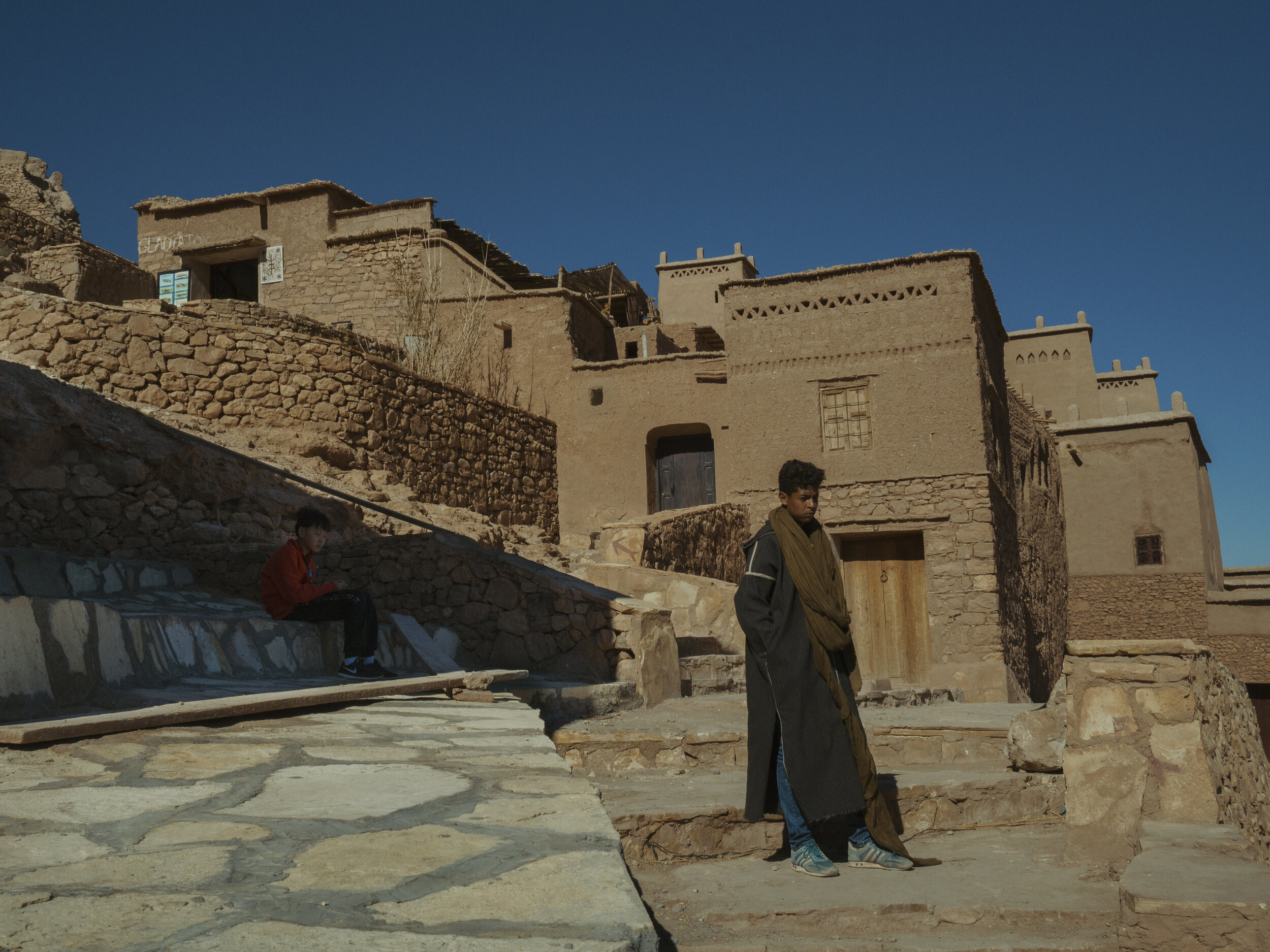 Two young men in a Moroccan village; one stands on stone steps, the other sits, with earthen houses and clear blue sky in the background.