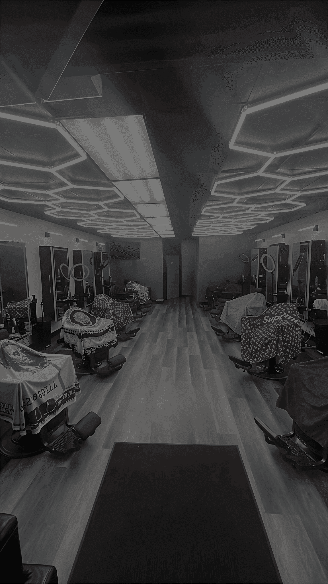 A modern barbershop interior with several barber chairs lined up along both walls, each covered with colorful capes. The space features large mirrors, ring lights, and sleek black and gray finishes. The ceiling is decorated with geometric LED lighting, creating a stylish and contemporary atmosphere. The floor is wood-patterned vinyl, and the shop appears clean and organized.