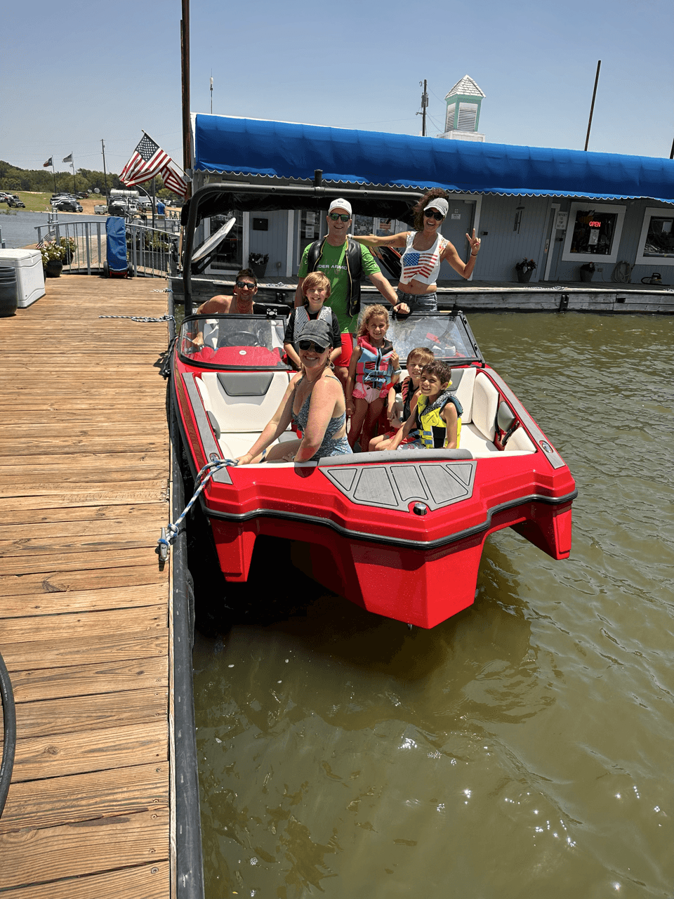 A red motorboat with a group of people, including children wearing life jackets, is docked at a wooden pier under a clear blue sky, with an American flag flying in the background.