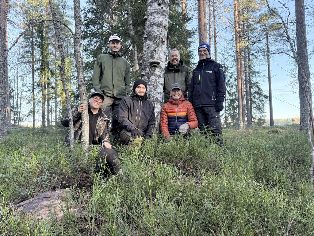 A young person with curly hair smiles at the camera while sitting in front of a laptop against a dark background.