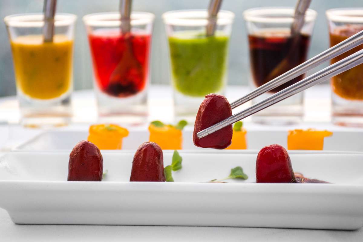 A pair of tongs holds a red fruit over a white rectangular plate containing several pieces of fruit and garnish.