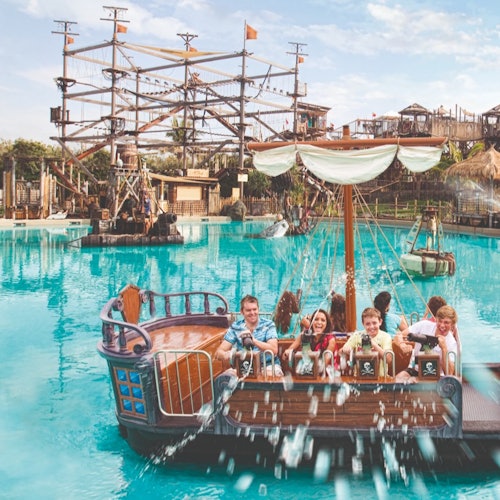 Mensen genieten van een tocht op een piratenthema-boot in een waterpark, met een touwparcoursstructuur zichtbaar op de achtergrond.