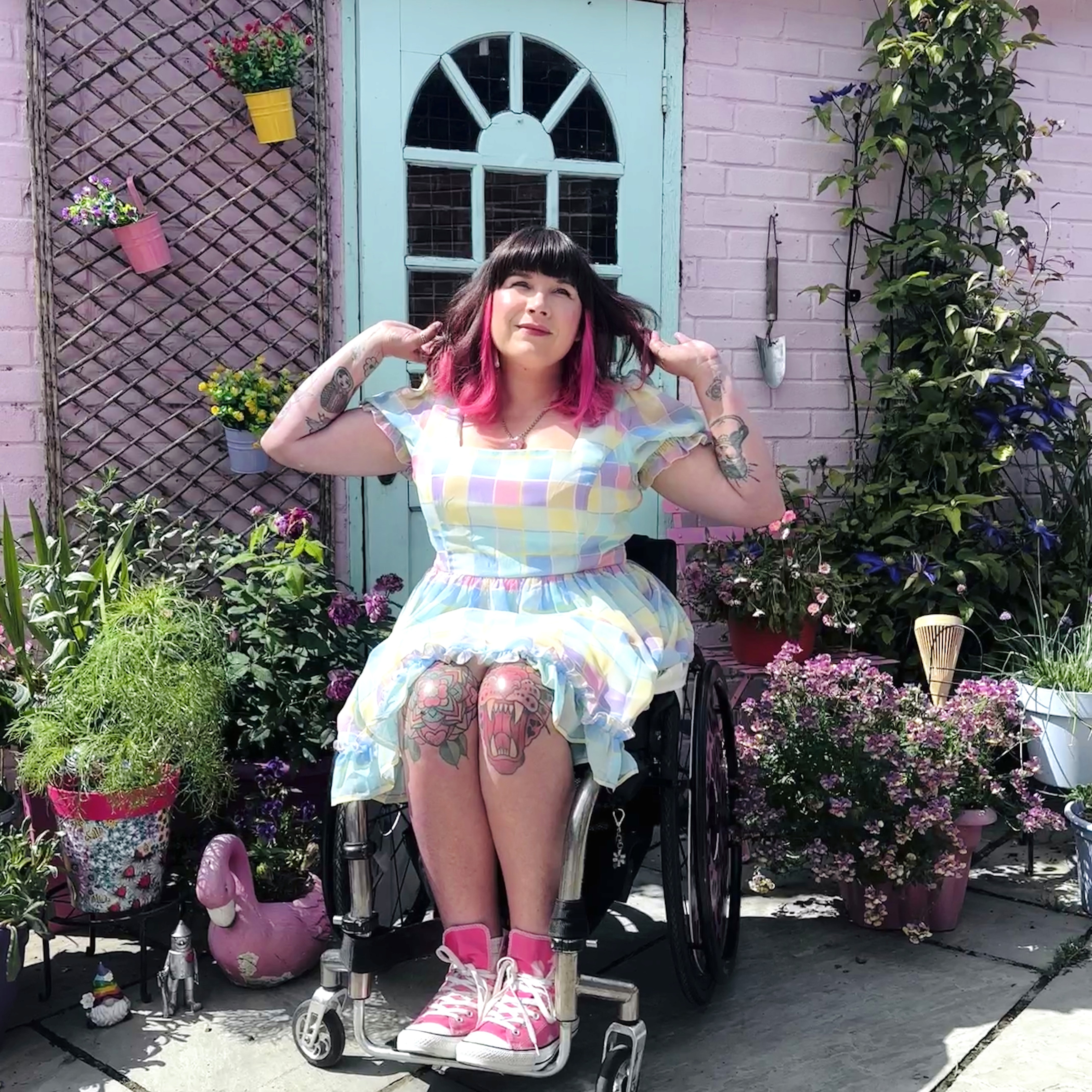 Nina, a wheelchair user with black and pink hair, is posing in front of her front door wearing a pastel checkered dress and pink high top converse. The door is mint against a pink wall and there are lots of potted plants all around.