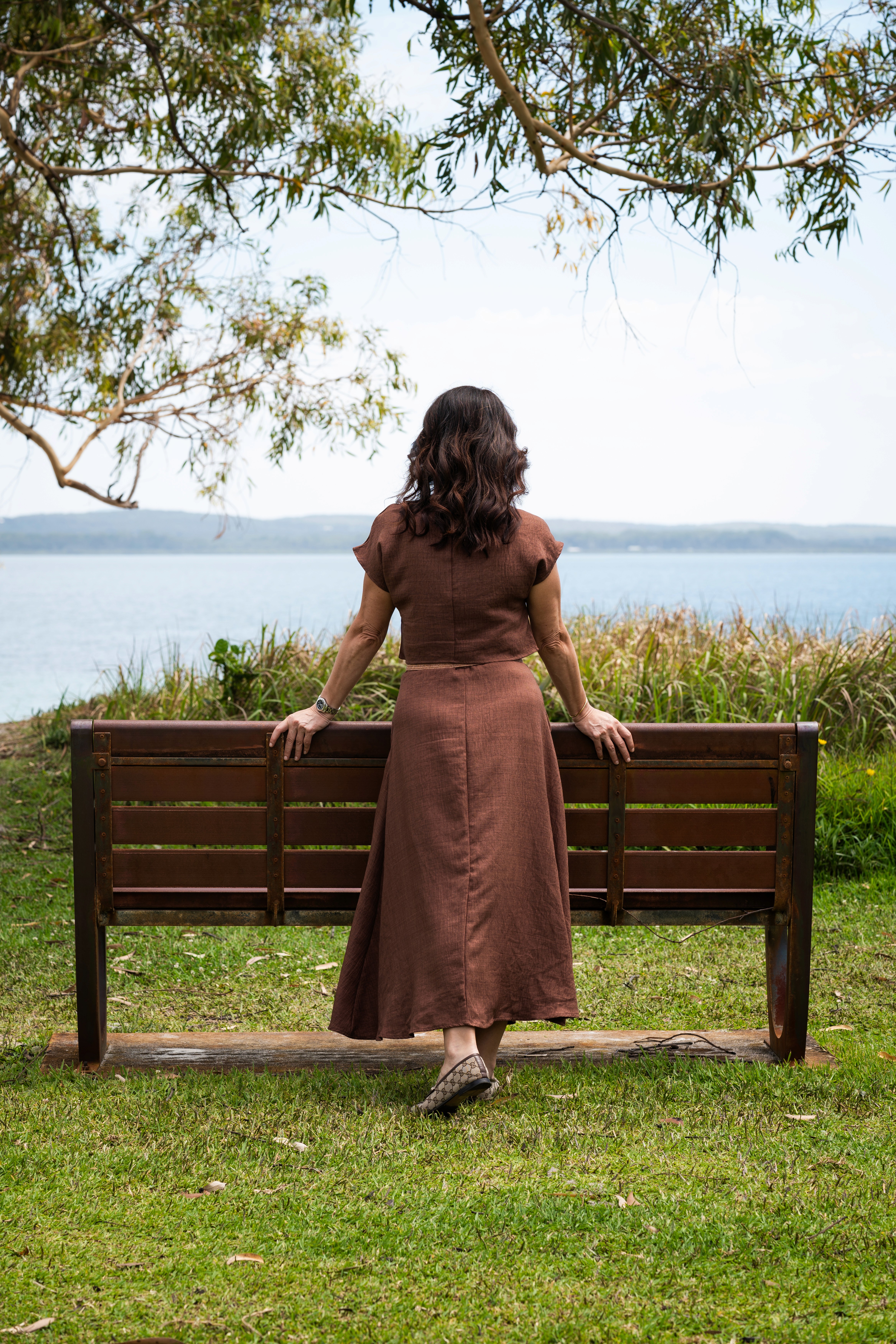 Dr. Bonnie Hawthorne in a brown dress stands by a wooden bench, looking out at a scenic lakeside view.