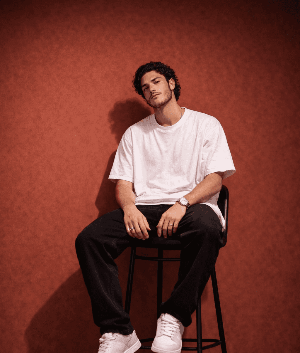 Young male designer sitting on a high chair against a warm terracotta studio background, wearing oversized white t-shirt, black pants and white sneakers