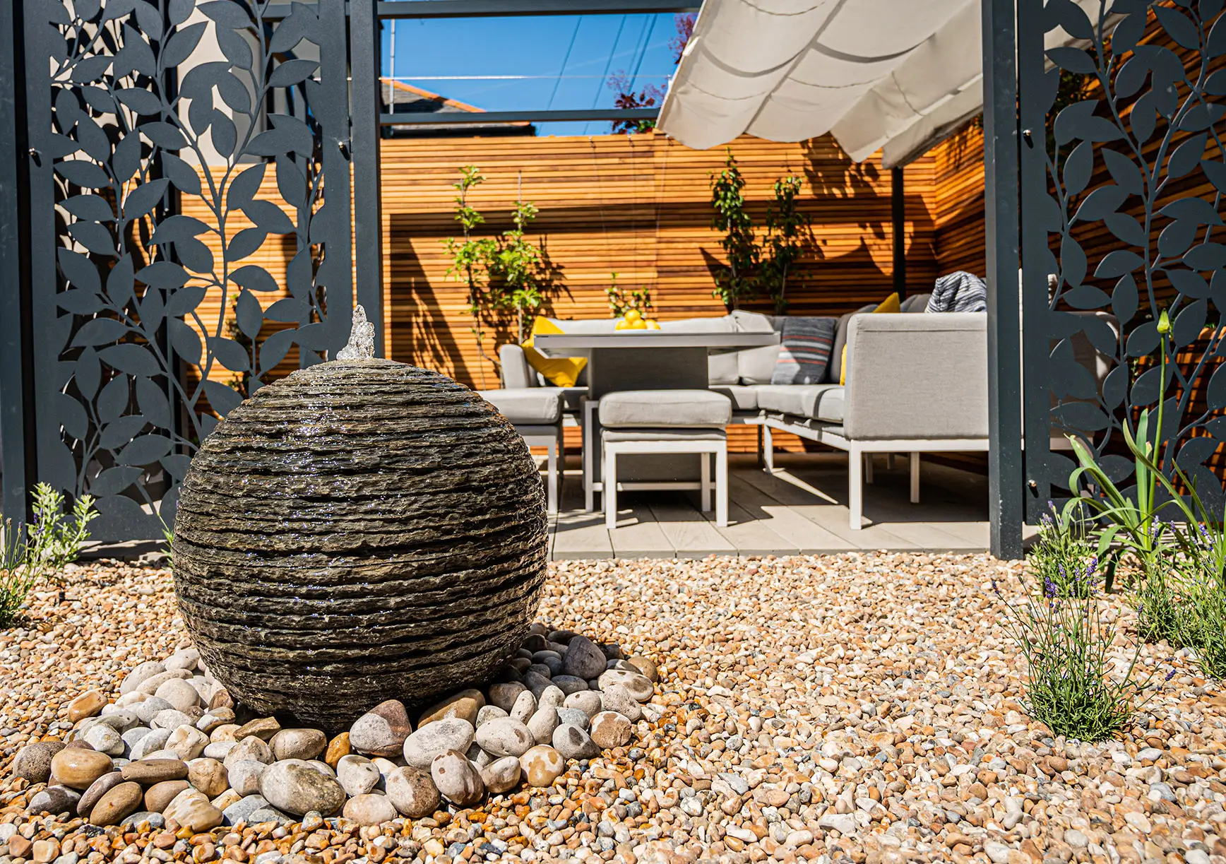 A round stone sculpture in a landscaped garden with seating and wooden walls in the background, under blue sky.