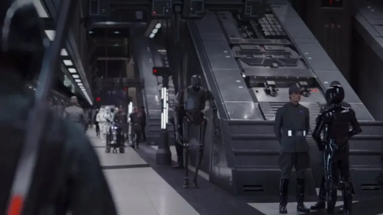 A futuristic, dimly-lit corridor features uniformed personnel and various droids engaged in discussion, surrounded by advanced technology and control panels in a high-tech facility.