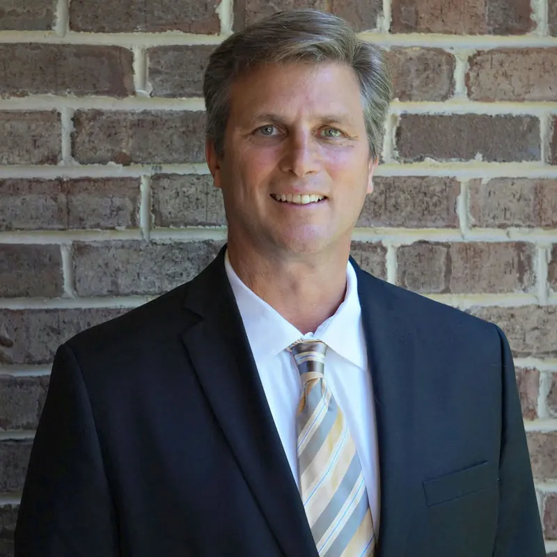 Fotografía de un hombre de mediana edad con traje oscuro y corbata, con expresión seria y profesional, representando el perfil de un directivo senior en el sector de la construcción.