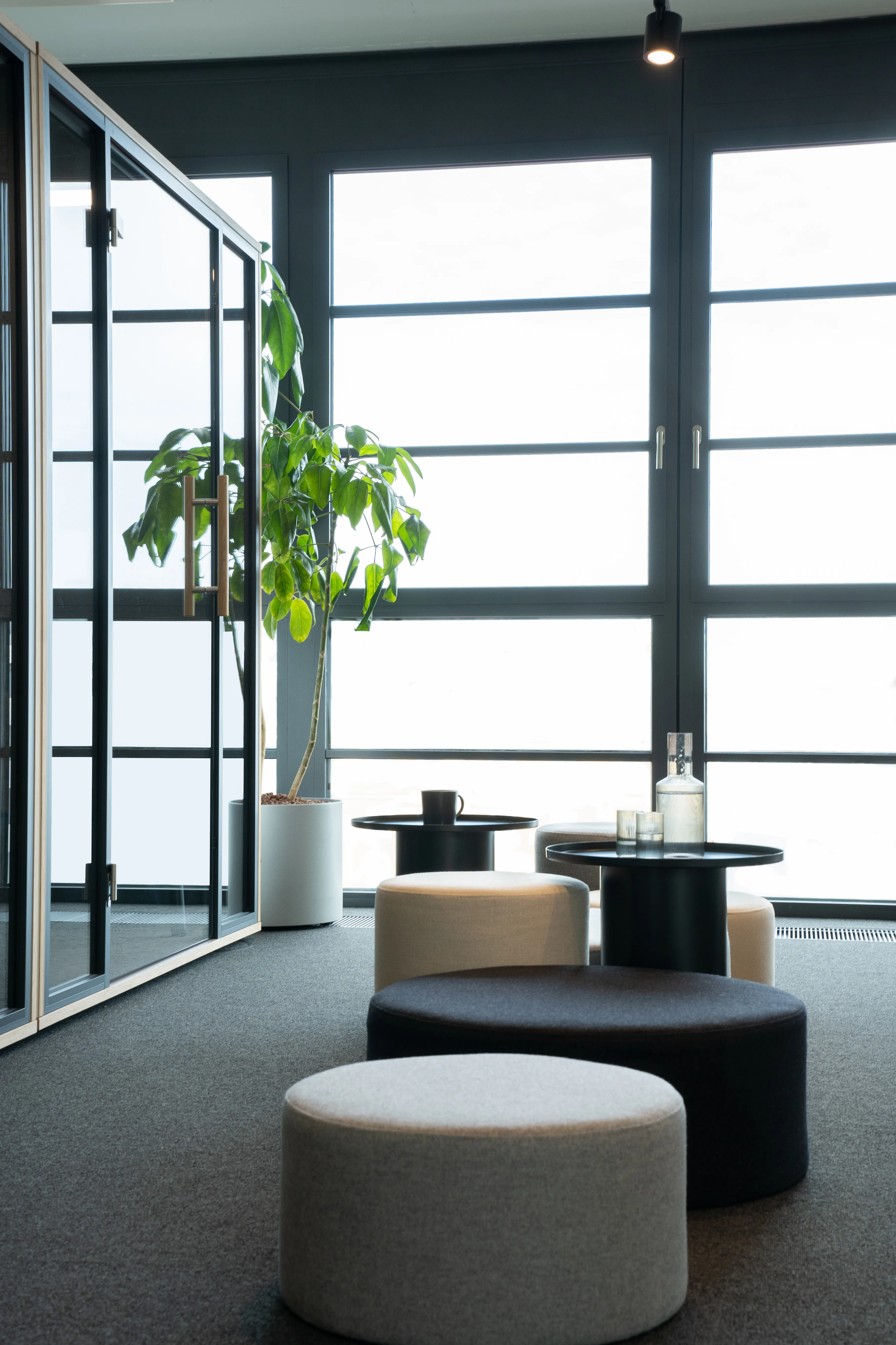 Lounge corner at AI START-UP MUNICH with black-framed glass partitions, a tall potted plant, black round tables and fabric poufs in cream and black on dark carpet.
