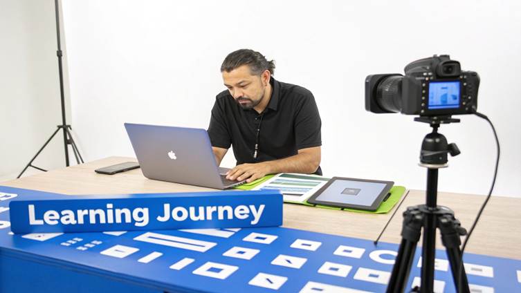 A man sits at a desk, typing on a laptop, with a camera recording in a studio setup for a learning journey.