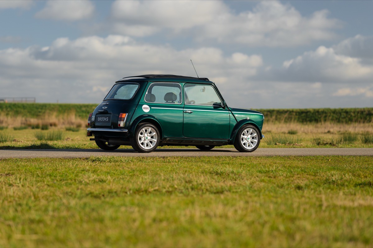 A classic green car driving on a rural road with fields and a blue sky in the background.