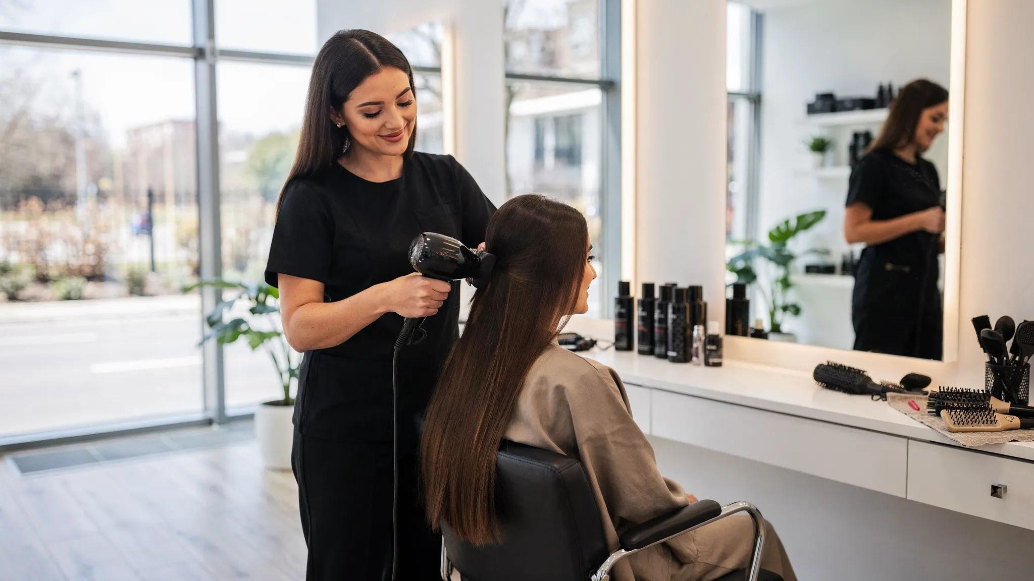 A bright, modern hair salon interior with a stylist finishing a client’s hair at the mirror, showing glossy, healthy-looking hair and neatly arranged professional products in the background.