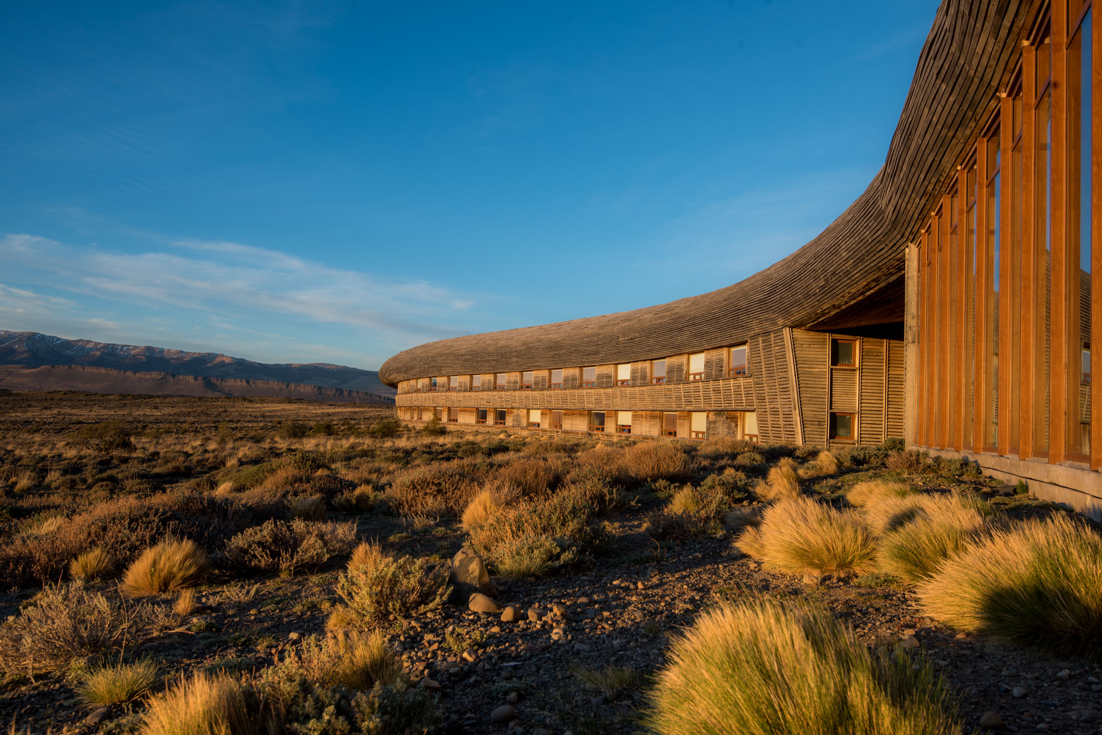 A modern wooden building with a curved roof and large glass windows sits in a dry, grassy field under a blue sky.